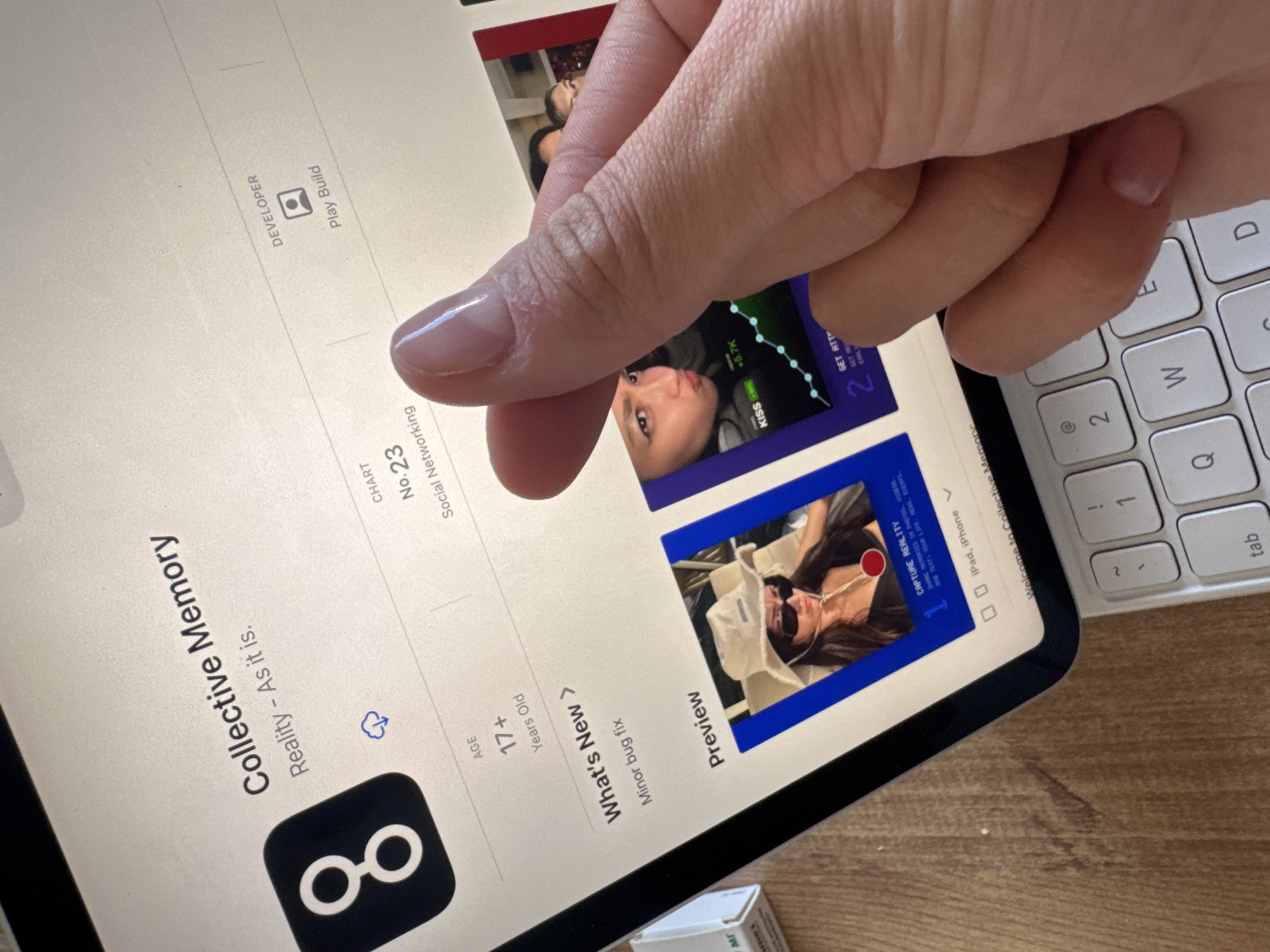 An overhead shot captures a hand holding an iPad, which displays an App Store page for an application named "Collective Memory." The hand, with neatly manicured, light pink fingernails, partially covers the screen content. The iPad rests on a light-colored wooden surface, with a white Magic Keyboard visible to its right.
The iPad screen prominently features the "Collective Memory" app, described as "Reality - As it is." Its icon is a black square with a white infinity-like symbol. Key app details include an age rating of "17+ Years Old," a "Minor bug fix" noted under "What's New," and its classification as "No.23 Social Networking." The developer is listed as "Play Build."
Two app preview images are visible. The first shows a person with dark hair, seemingly pouting or making a kissing face, overlaid with a green line graph and text like "+5.7K KISS." The second preview depicts a person, likely female, wearing sunglasses and a hat, with text that reads "CAPTURE REALITY SHARE MEMORIES IN PHOTOS, NO TEXTS, VLOG LIFE, EVENTS." The screen also indicates compatibility with "iPad, iPhone" and a "Welcome to Collective Memory" banner.
Part of the white keyboard on the right shows keys such as "tab," "Q," "W," "E," "D," "A," "2," "@," and "!." A small white box with faint text, possibly "мг," is discernible in the bottom right corner of the frame. The lighting suggests an indoor, daytime setting.