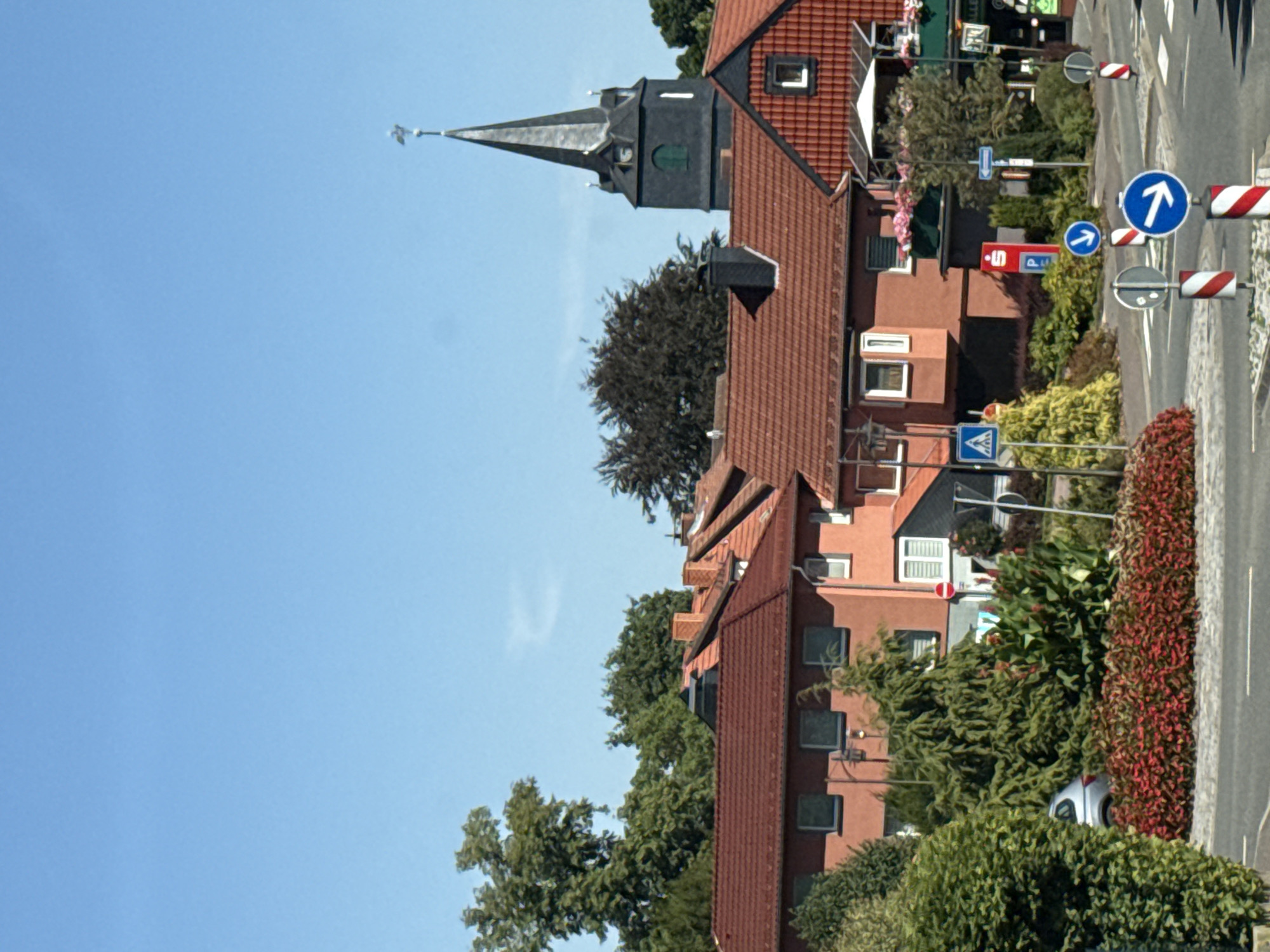 The image shows a clear blue sky above a street scene in Bad Sachsa, Germany. In the foreground, a paved road curves to the left, bordered by a grey curb and a band of low-lying red foliage. Behind the foliage, a silver car is parked next to a large evergreen tree. Further back, several multi-story buildings with red tile roofs are visible, some with brick or stucco facades in shades of orange and tan. One building has a prominent dark grey church tower with a steeple topped by a weather vane. Various traffic signs, including directional arrows and a pedestrian crossing sign, are positioned along the roadside. The scene appears to be set in a residential or commercial area with lush greenery and well-maintained buildings.