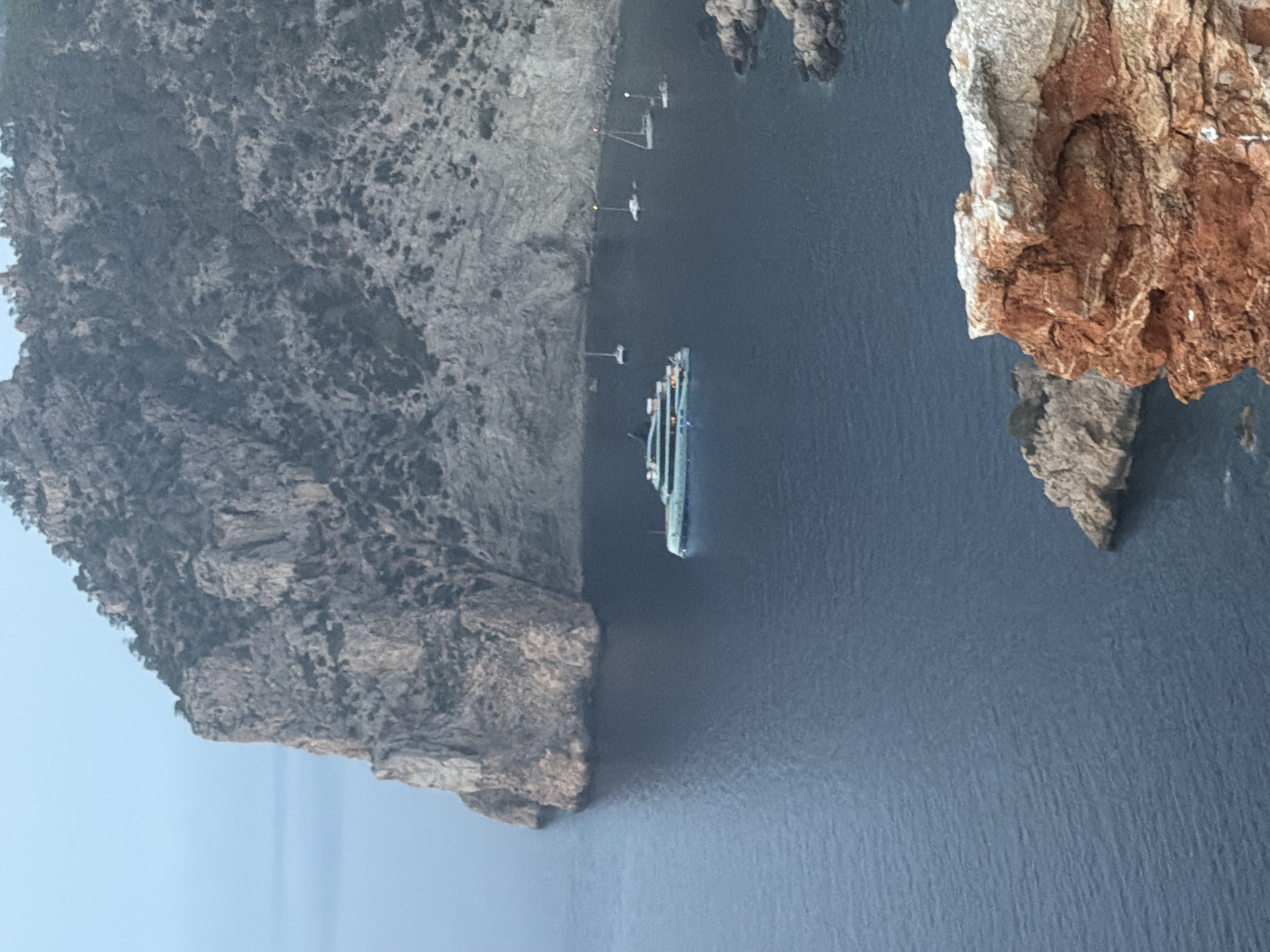 The image captures a coastal landscape dominated by a large, rugged cliff face. The cliff is covered in sparse vegetation and appears to be made of grey rock. At the base of the cliff, the dark blue sea laps against the shore. A modern, light blue yacht with multiple decks and illuminated windows is anchored in the calm water near the cliff. Several smaller sailboats are also visible in the bay, some with their sails down. In the foreground, reddish-brown rocks form a rocky outcrop overlooking the sea. The sky is overcast and a pale blue, suggesting either early morning or late evening. The scene appears to be a scenic bay, likely a popular anchorage or mooring spot. While the image is described as being in Sant Antoni de Portmany, Spain, the specific location within the city is not discernible from the visual cues alone. There are no people visible in the image.