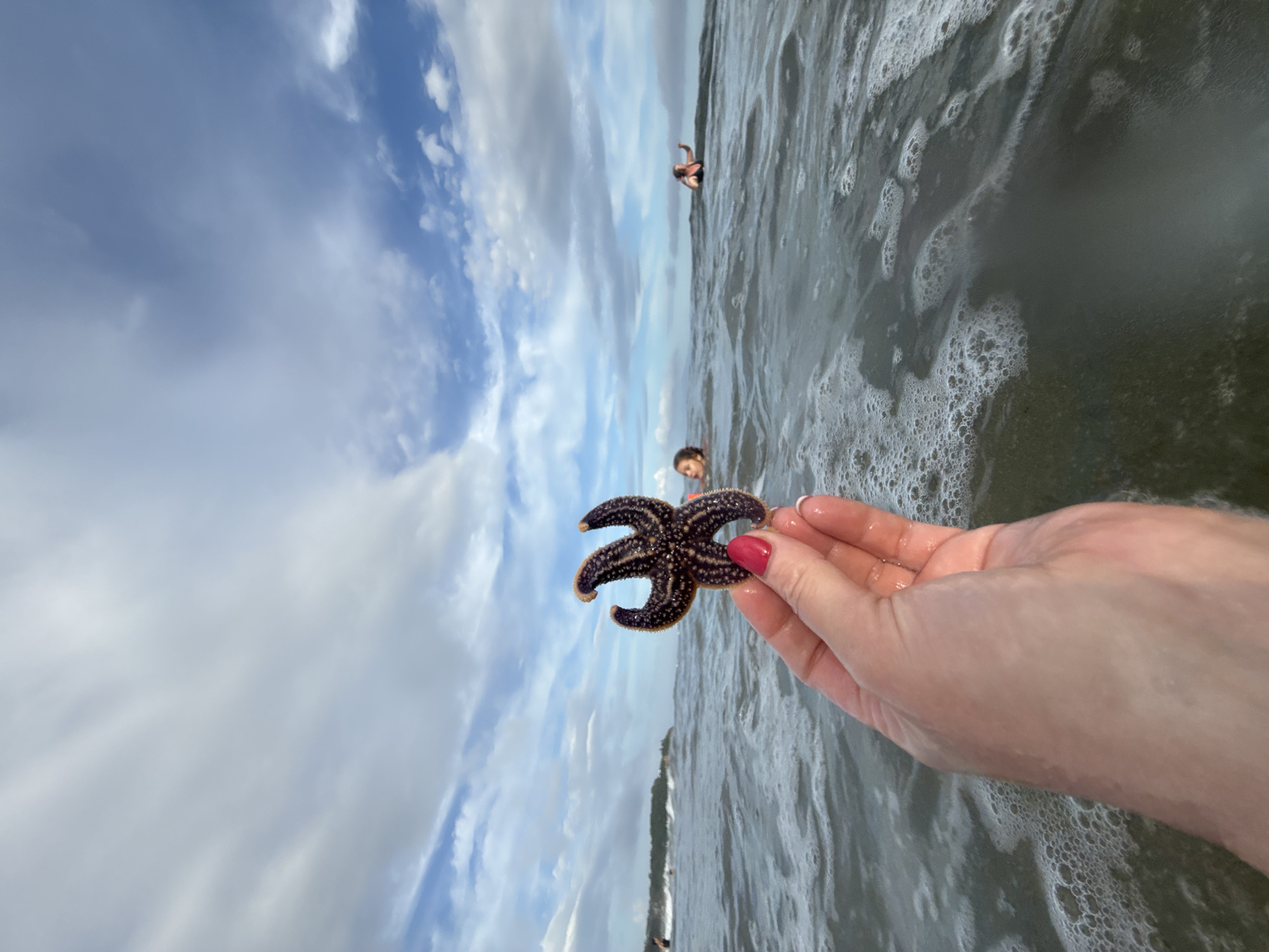 The image shows a close-up of a person's hand holding a dark-colored starfish with prominent knobs on its arms. The hand has bright red nail polish on one finger. The starfish is held in front of a slightly out-of-focus ocean with waves and white foam. In the background, several people are visible in the water, with their heads above the surface. One person on the right appears to be reaching their arm up. The sky is cloudy with patches of blue. The location is not identifiable as Vladivostok, Russia.