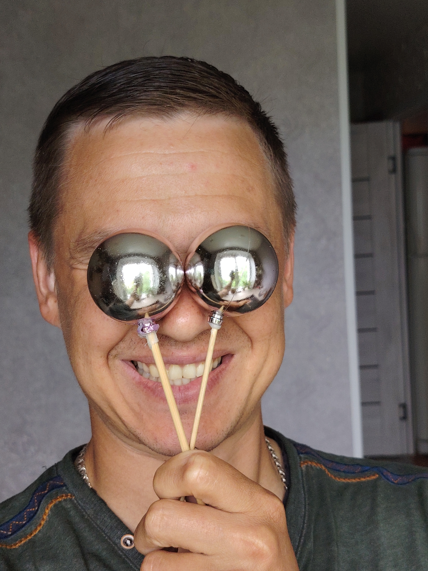 A male individual is captured in a mid-torso to headshot, smiling broadly, and playfully holding two spherical, highly reflective objects on sticks in front of his eyes, obscuring them. The man has short, dark hair, light-to-medium skin tone, and is wearing a dark olive-green long-sleeved shirt with embroidered patterns in orange and blue along the shoulder seams. A silver-colored curb chain necklace is visible around his neck. His expression is a wide, open-mouthed smile, revealing a full set of teeth. His right hand (viewer's left) holds the two light-colored wooden sticks, which converge near his lower face. One stick features a purple-colored band securing the spherical object, while the other has a silver-colored metallic cap at the base of the sphere. The reflective surfaces of the spheres show distorted reflections of a bright outdoor environment, including what appears to be trees and sky. The background consists of a plain, light-gray wall. To the right of the subject, a vertical white trim, likely part of a doorframe or wall edge, is visible, beyond which darker, less distinct background elements can be seen. The scene is an indoor setting. There are no visual indicators within the image to specify a location in Chaikovsky, Russia.