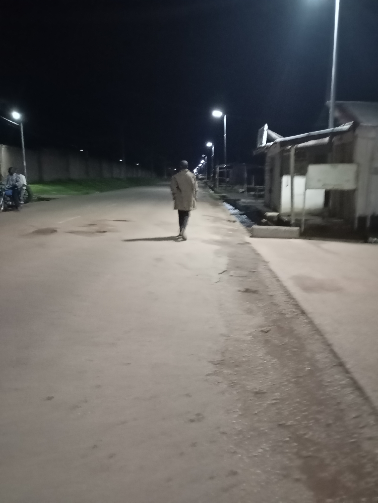 The image depicts a nighttime scene on a paved road in Shendam, Nigeria. A single individual, appearing to be a male, is walking away from the camera down the middle of the road, wearing a light-colored coat and dark trousers. To the left of the road, a person is seated on a motorcycle, with another motorcycle visible behind it. Streetlights illuminate the road, casting long shadows. Buildings and a wall line the right and left sides of the road, respectively. The overall atmosphere is dim and quiet.