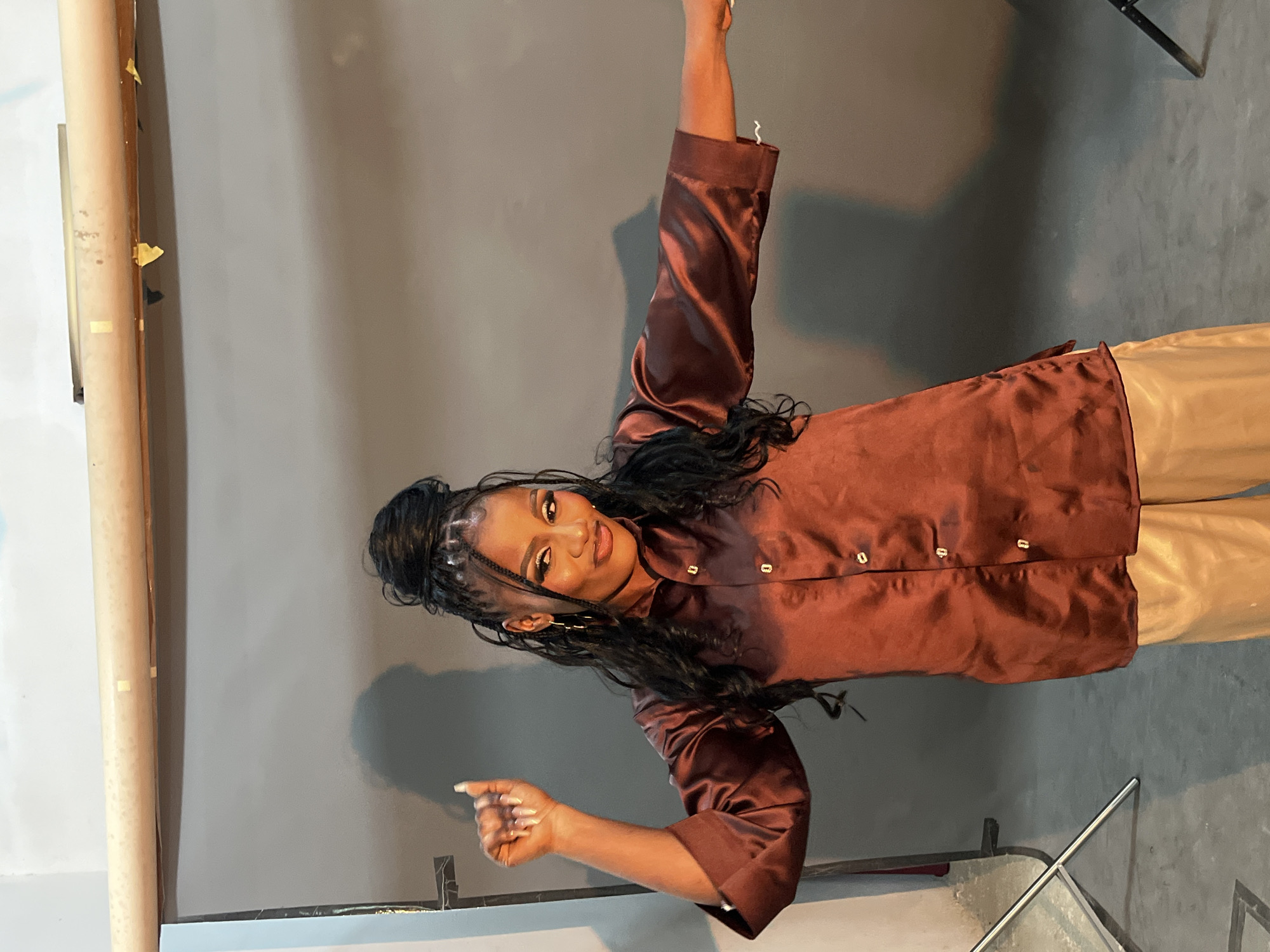 A young Black woman with long, dark braided hair styled in a high bun is posing in a studio setting. She is wearing a satin, long-sleeved, dark red collared shirt with buttons down the front and light tan pants. She has dramatic eye makeup, hoop earrings, and her left arm is raised with her fist clenched. Her right arm is extended to the side. The background consists of a gray studio backdrop, with a light-colored bar and some tape visible at the top. Lighting equipment stands are partially visible on the left and right sides of the frame. The floor appears to be a dark gray concrete. There is no indication of the location being in Gura Top, Nigeria, or any specific city.
