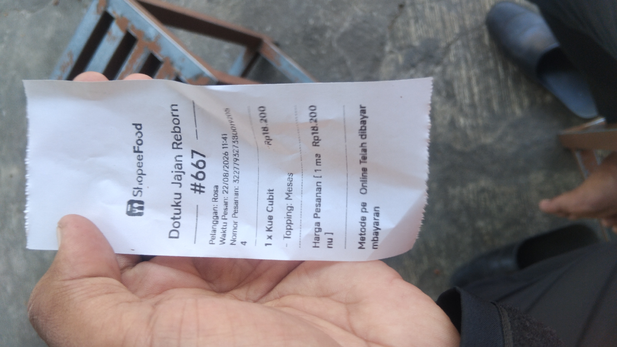 The image shows a hand holding a ShopeeFood receipt. The receipt is for "Dotoku Jajan Reborn" and has an order number "#667". It lists the customer as "Rosa", the order time as "22/08/2026 11:41", and the order number as "3227793275300192054". The item ordered is "1 x Kue Cubit" with "Topping: Meses". The total price of the order is Rp18.200. The payment method is "Online Telah dibayar". The background shows a concrete floor and a part of a metallic chair. The image is taken from a slightly overhead angle. There are no discernible landmarks to place this image within the city of Proyonangg, Indonesia.