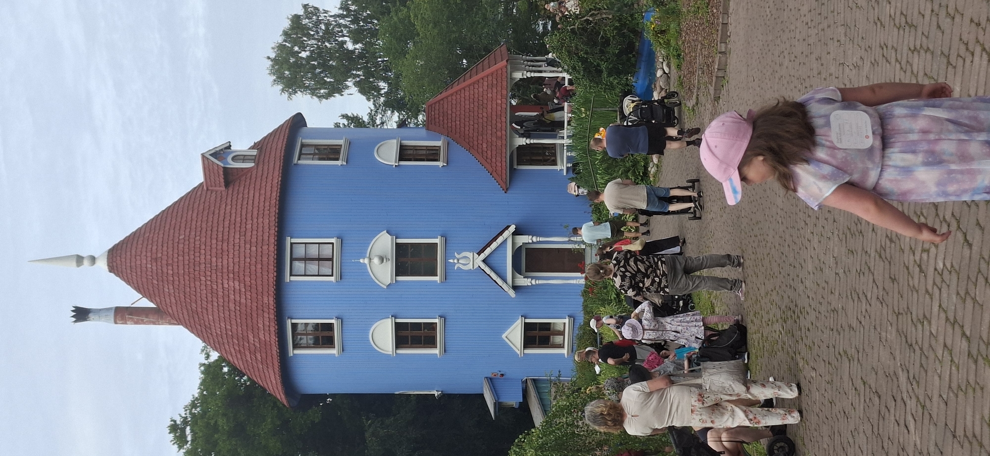The image depicts the Moomin House, a fictional residence from Tove Jansson's Moomin books, located at Moominworld in Naantali, Finland. The iconic blue, multi-story house features a distinctive red, conical roof and a tall chimney. Several people are present in the foreground and around the house, including adults and children. Some are walking, others appear to be observing the house or interacting with each other. The environment is a park-like setting with paved pathways, lush greenery, and trees. The sky is overcast, suggesting a cloudy day. A young girl in a pink cap and a colorful dress is prominently featured in the foreground, walking away from the camera.