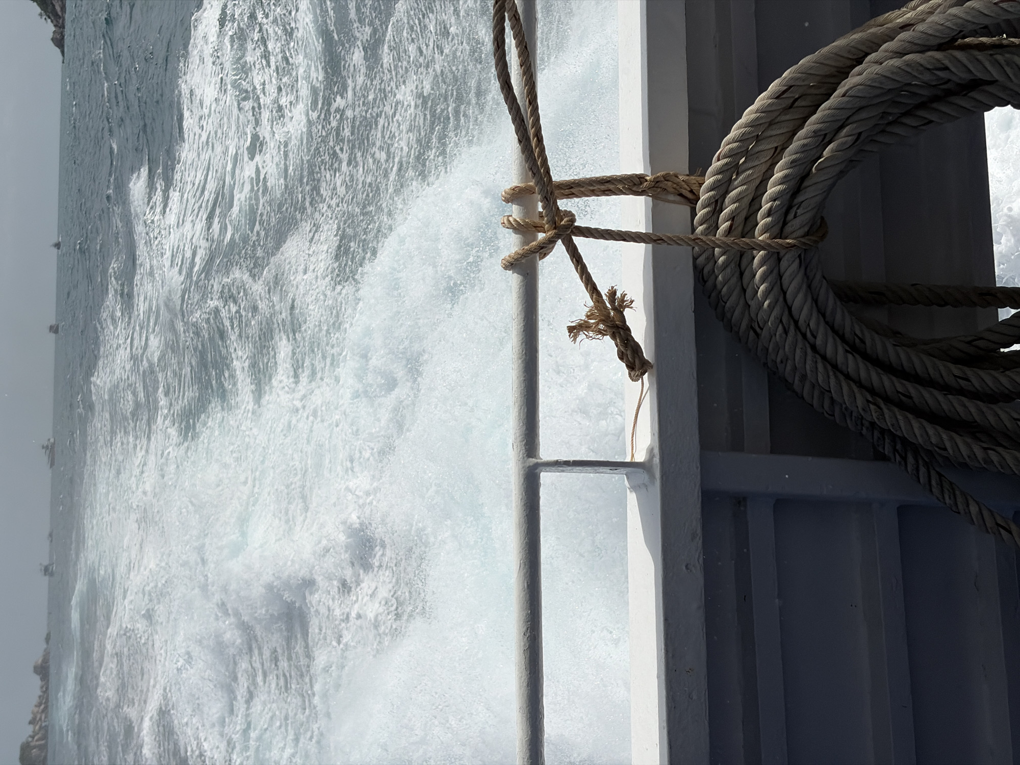 A boat is seen from the stern, moving through rough, choppy, turquoise water with a foamy wake. In the distance, several boats are visible on the horizon, along with rocky islands and a hazy sky. A thick rope is coiled and hanging from a railing on the right side of the boat, secured by a knot. The railing is white and appears to be made of metal. The deck of the boat is visible in the foreground, with gray siding. The image was taken from the deck of a moving boat, suggesting a ferry or tour boat operating in the waters around Ko Pha-ngan District, Thailand.