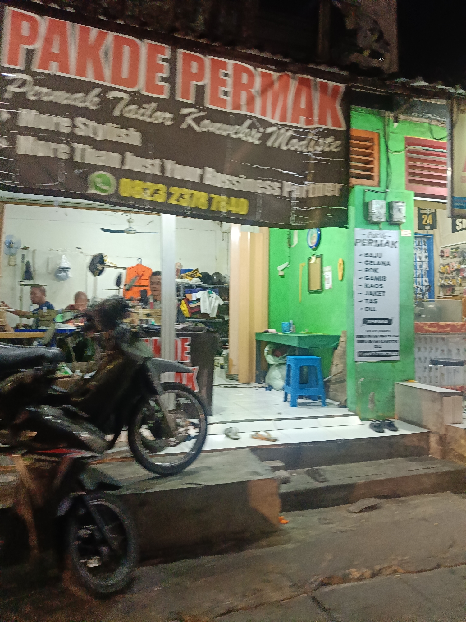 The image shows a storefront in Surabaya, Indonesia, with a large banner that reads "PAKDE PERMAK" in red lettering. Below this, smaller text states "Permak Tailor Konveksi Modiste" and then "More Stylish" and "More Than Just Your Business Partner." A WhatsApp logo and a phone number, "0823 2378 7840," are also visible on the banner.
The storefront itself appears to be a tailor shop, with mannequins and clothing racks visible inside. Three men are working inside the shop, one of whom appears to be at a sewing machine. A motorcycle is parked in front of the shop, partially obscuring the entrance. The shop is painted green and has a light-colored tiled floor. A blue stool is placed near the entrance. A sign in Indonesian lists clothing items: "BAJU," "CELANA," "ROK," "GAMIS," "KAOS," "JAKET," and "TAS," along with the word "DLL" (etc.).
The lighting suggests it is nighttime or dusk. The overall impression is of a small, independent tailor shop operating in a commercial or residential area.