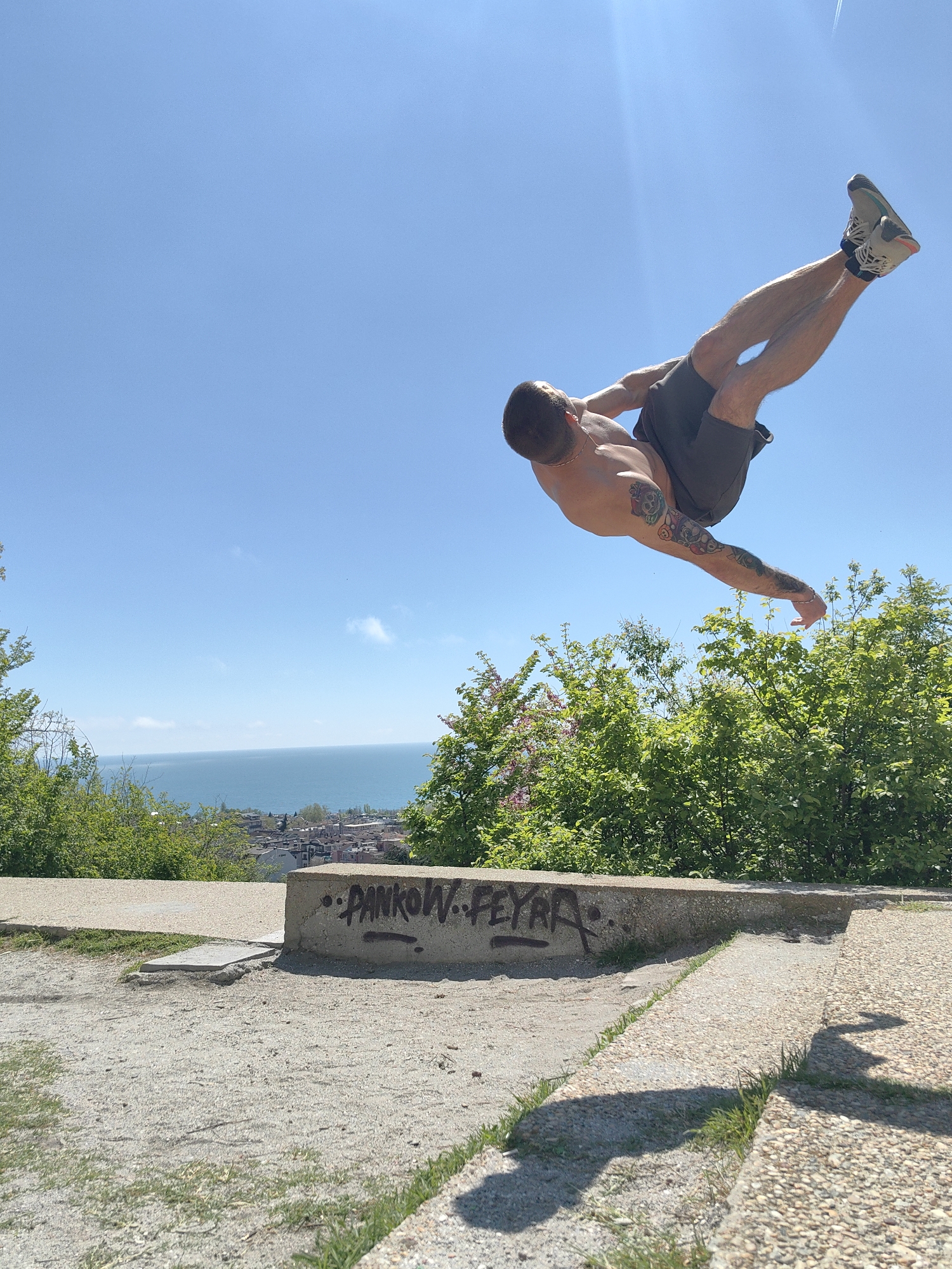 A man is performing a parkour move mid-air in Varna, Bulgaria. He is shirtless, wearing shorts and sneakers, and has tattoos on his arm. He is positioned above a low concrete wall with graffiti on it, in a park setting with green trees and a view of the Black Sea and the city of Varna in the background under a clear blue sky. The ground in the foreground is a mix of dirt and concrete.