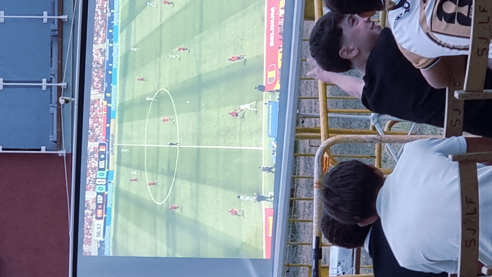 The image captures a projection of a soccer match on a large screen, likely outdoors, given the surrounding environment. The screen displays a game between "ESP" (Spain) and "BEL" (Belgium), with the score at 0-0 in the 6th minute and 57 seconds. Several players in red and white uniforms are visible on the green field. Below the screen, in the foreground, are several people seated in chairs, facing away from the camera. The chairs have markings that read "SJ. LF" and "SJ". In the background, yellow metal fencing is visible, suggesting a public viewing area or a casual event. The overall setting appears to be an outdoor venue in Sant Joan Les Fonts, Spain, where a community might gather to watch a sporting event.
