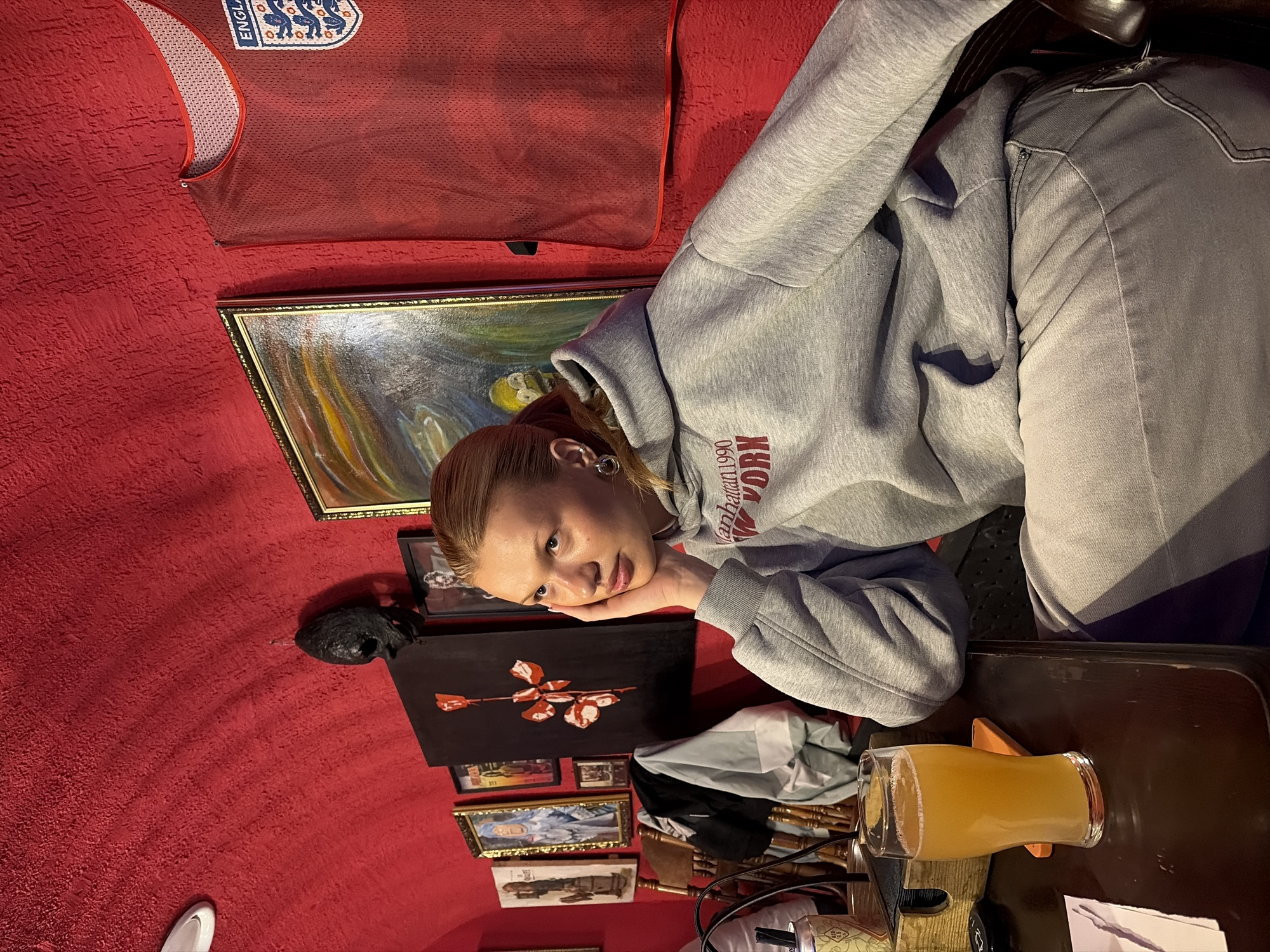A young person is seated at a table in a room in Chaikovsky, Russia. They are wearing a gray hooded sweatshirt with "Manhattan, New York 1990" printed on it and gray jeans. On the red wall behind them, there is a red jersey with the England emblem, an oil painting with a gold frame, a black painted canvas with a white and red rose, and several small framed photos and artwork. On the table, there is a glass of beer. The room has a red wall and ceiling.