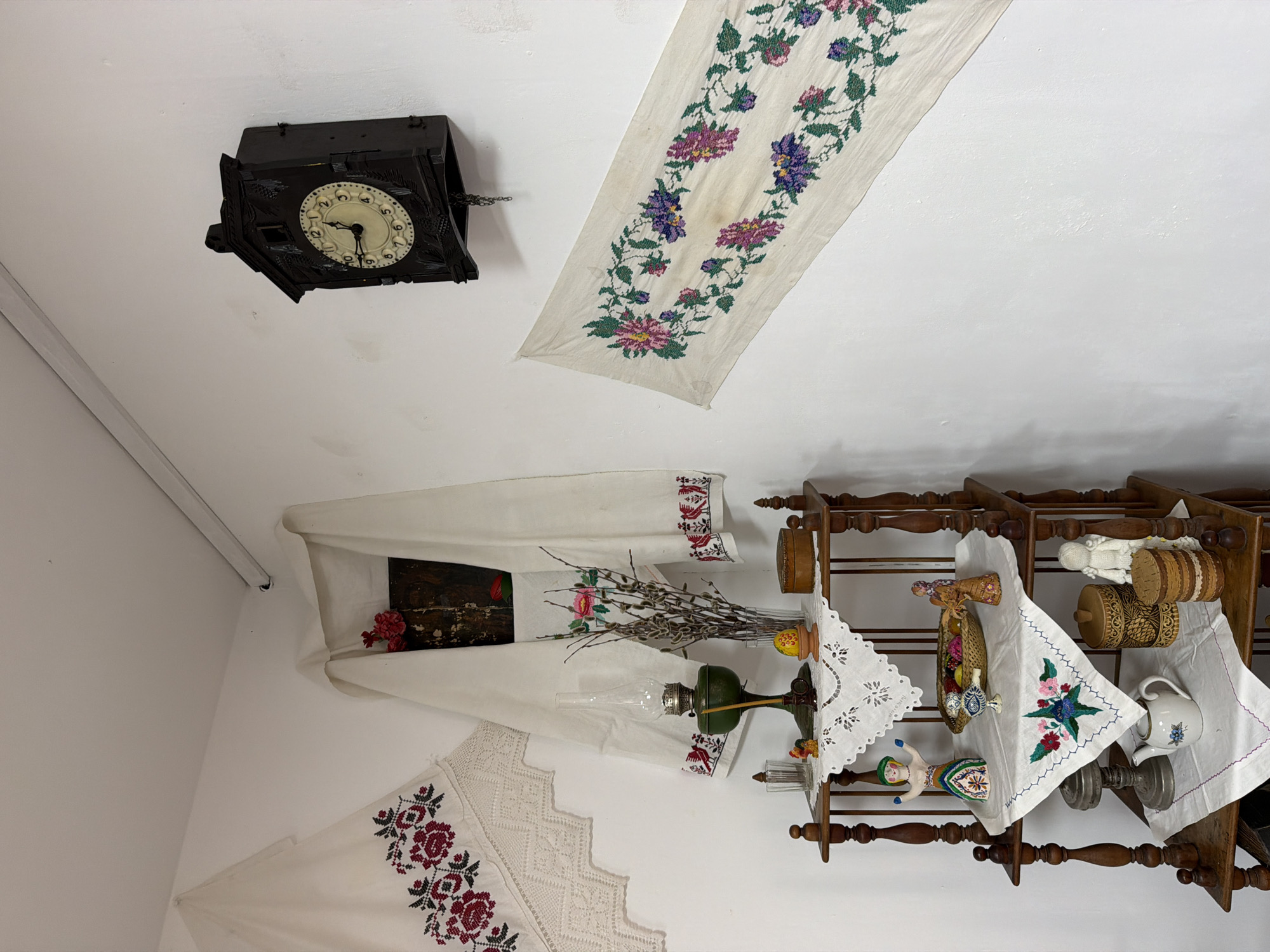 This is an indoor scene in Ust'-Kabyrza, Russia, likely a room within a traditional dwelling or museum. The environment is sparsely decorated with white walls and a wooden shelf unit. On the walls are two pieces of embroidered fabric: one with red floral patterns hangs to the left, and another with colorful floral patterns runs diagonally across the right. A dark wooden cuckoo clock hangs on the wall above the embroidered fabric on the right.
The wooden shelf unit displays various decorative objects. The top shelf holds an oil lamp with a green base and glass chimney, a candle, a small egg in a stand, a small wooden box, and a glass container with a rooster figurine. The second shelf features a doll in a colorful dress, a basket filled with colorful objects, a small bird figurine, and another figurine resembling an angel. The third shelf displays a metal object, a teapot with a floral design, a round wooden container with a patterned surface, and a standing figure made of what appears to be white yarn or fabric. A piece of embroidered fabric with a floral design is draped over the third shelf.