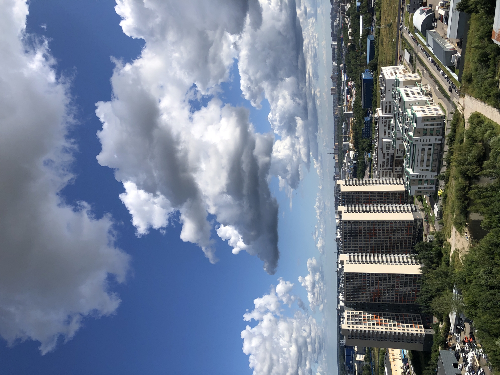 The image depicts an aerial view of a modern urban landscape under a partly cloudy blue sky. In the foreground, a cluster of tall, multi-story residential buildings dominates the scene. These buildings have a uniform architectural style, characterized by light-colored facades and numerous windows, some of which appear to have orange accents. Adjacent to these taller structures, there are lower, more horizontally oriented buildings with green-capped roofs.
A network of roads and parking lots is visible, with cars parked along some of them and in designated areas. Sparse green vegetation, including trees and grassy areas, is interspersed throughout the built environment, with a small body of water present to the left of the foreground buildings. In the midground and background, more city structures and industrial buildings can be seen, along with a distant treeline. The sky is filled with large, fluffy cumulus clouds, some of which are dark and shadowed, suggesting dynamic weather conditions. The overall impression is of a developed urban area on a bright, albeit cloudy, day.