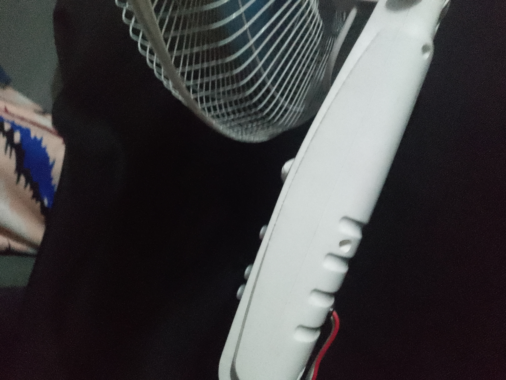 A close-up, slightly blurry, waist-level shot shows a white electric fan and a dark background. The fan's grill is made of silver metal wires and is partially visible on the left. A white plastic casing runs vertically down the right side of the frame, with several indentations or buttons visible. A thin red wire is attached to the bottom of the white casing. To the left of the fan, a piece of fabric with a blue and black zig-zag pattern is visible against the dark background. The environment appears to be indoors, with no specific urban landmarks identifiable.