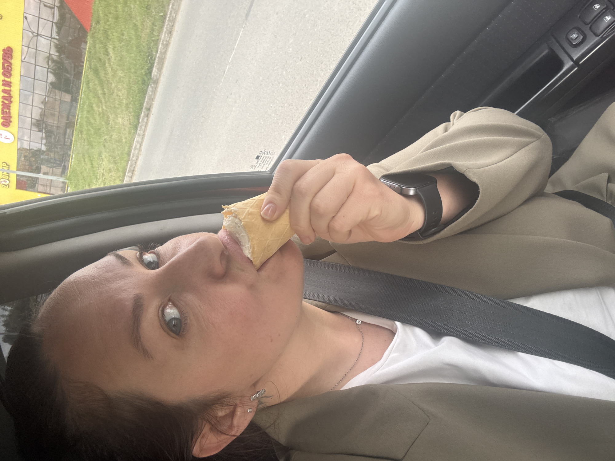A woman, appearing to be in her 30s-40s with dark brown hair, fair skin, and light blue eyes, is seated inside a vehicle, looking directly towards the viewer. She is wearing a khaki-colored blazer over a white crew-neck t-shirt. A black automotive seatbelt is diagonally secured across her chest. Her left ear displays two small silver stud earrings and a small silver hoop earring. A delicate silver chain necklace with a single small, round diamond pendant is visible on her collarbone. She is holding a waffle cone containing white ice cream to her mouth, actively eating it. Her left wrist bears a dark-colored smartwatch. The vehicle's dark plastic door panel, complete with power window controls, is visible to her right. Outside the window, a paved road or shoulder is present, bordered by a strip of green grass. In the background, a modern building with a facade of large, reflective glass windows is visible, showing reflections of trees. Above the building, a yellow sign displays Russian text in red capital letters, partially visible as "ZOOM P" (likely "ZOOM.IP") and clearly "ОДЕЖДА И ОБУВЬ" (meaning "CLOTHES AND SHOES"), accompanied by a red icon resembling a clothes hanger. This sign corresponds to a known retail store in Chaikovsky, Russia, specifically identifying the location as the city of Chaikovsky, Russia.
