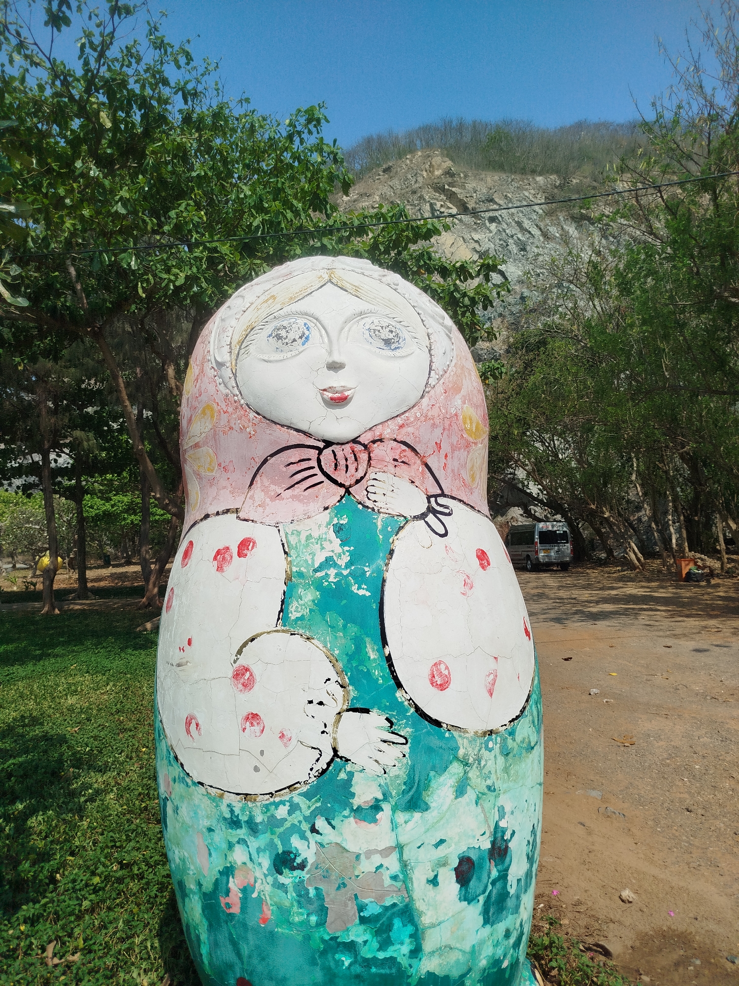 The image shows a large, weathered, painted Matryoshka doll standing in what appears to be a park or roadside area. The doll's paint is cracked and peeling, revealing layers of pink, white, and teal. The doll has a simple, folk-art style face, with large blue eyes, a small red mouth, and a pink headscarf. Below the face, the doll's body is decorated with what appear to be additional, smaller dolls held in the larger doll's arms. The background features trees, a dirt road, and a hillside with exposed rock. A van is partially visible behind the doll. Given that there is a mountain in the background, this image may not have been taken in Ho Chi Minh City as it is mostly flat.