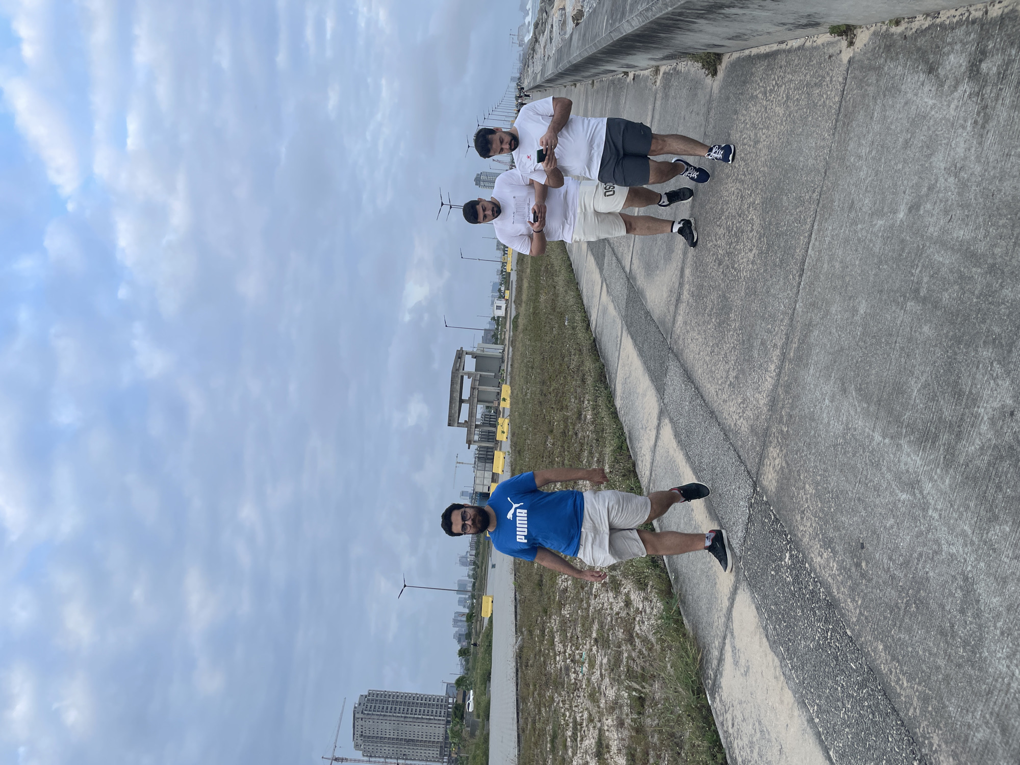 The image captures an overcast day with a cloudy sky over Inupa Kekere, Nigeria. A paved walkway runs along a concrete barrier, suggesting a waterfront promenade or similar coastal infrastructure. In the foreground, a man with dark hair and a beard, wearing a blue Puma t-shirt and light-colored shorts, walks towards the camera. Behind him, two other men in white t-shirts and shorts walk away from the camera, both looking at their mobile phones. The environment shows signs of ongoing development, with construction equipment and buildings visible in the background, including a tall skyscraper and a crane. The ground on the left side of the walkway is covered with dry grass and sand. The sea is visible to the right of the concrete barrier, with waves crashing against the shore.