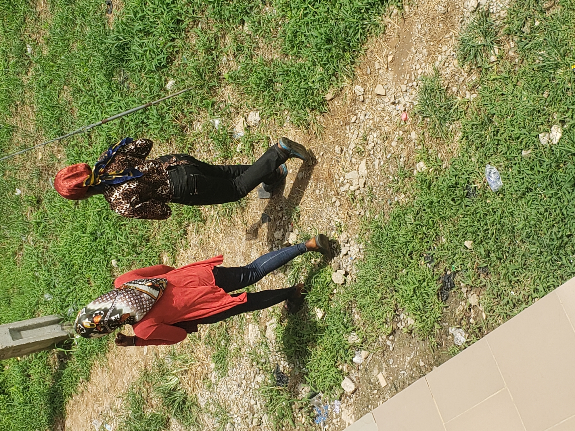 Two individuals, identified by their attire as women, are walking away from the camera on a dirt path bordered by green grass. The woman on the left wears a red long-sleeved top and dark blue jeans, her head covered with a patterned hijab. The woman on the right is dressed in a brown and black animal-print shirt, dark trousers, and a red headscarf. A metal pole stands vertically to the right of the individuals. The ground is a mixture of dirt, small stones, and patches of grass, with some scattered litter visible. The scene appears to be outdoors, possibly in an urban or semi-urban area given the presence of litter and the building visible on the left. The overall impression is a candid snapshot of everyday life.