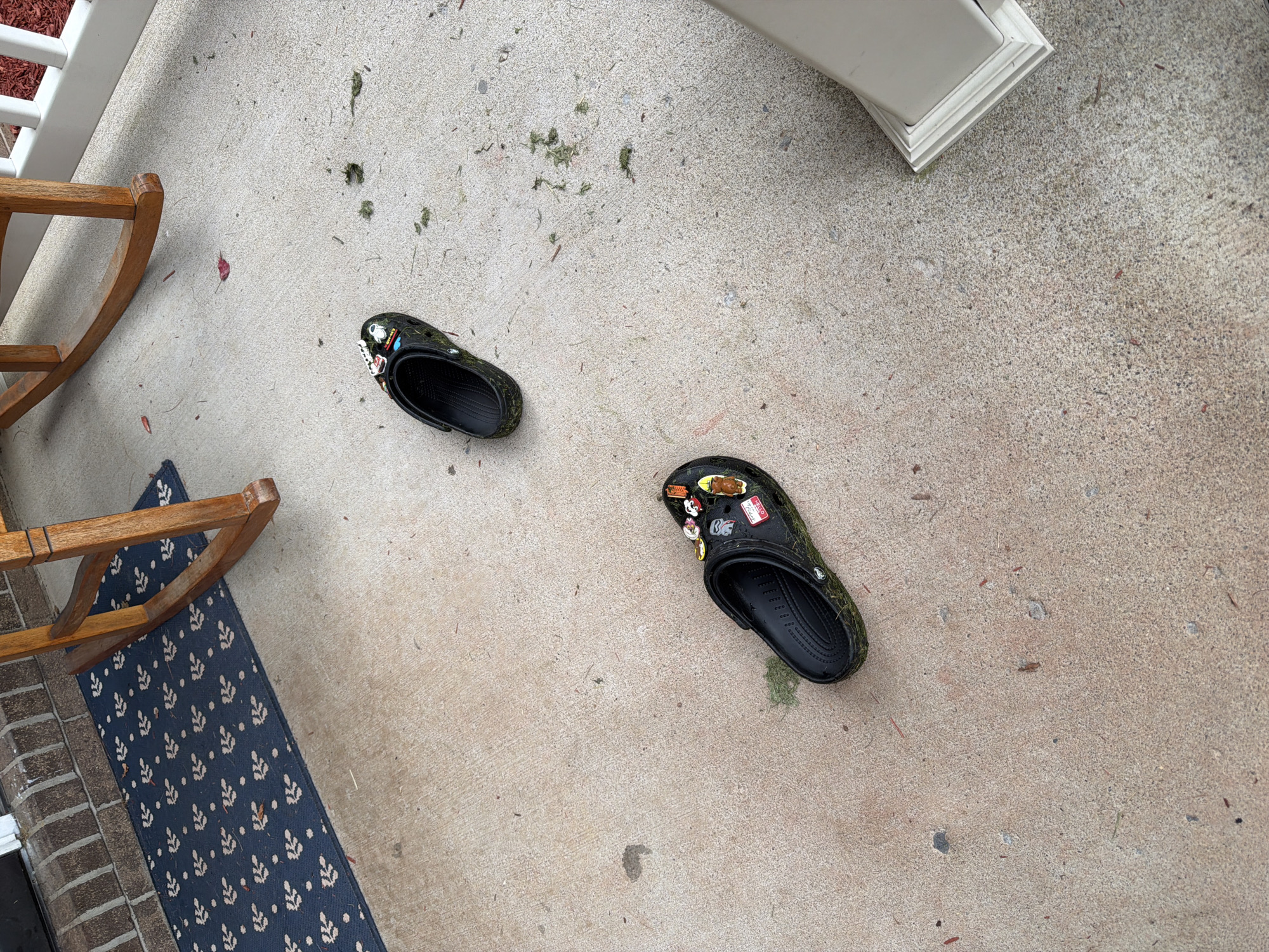 A pair of black Crocs, adorned with various decorative charms including what appears to be a cartoon astronaut and a depiction of a slice of pizza, are scattered on a concrete porch. One Croc is positioned closer to the center of the frame, while the other is further up and to the right. Both shoes are partially covered in green grass clippings, suggesting recent outdoor activity. The porch also has a dark blue welcome mat with a white leafy pattern, and a wooden rocking chair is partially visible in the upper portion of the image. Scattered on the porch are a few dry leaves and grass debris. The overall scene suggests an outdoor residential setting, possibly a front porch. The location is identified as Negley, United States.