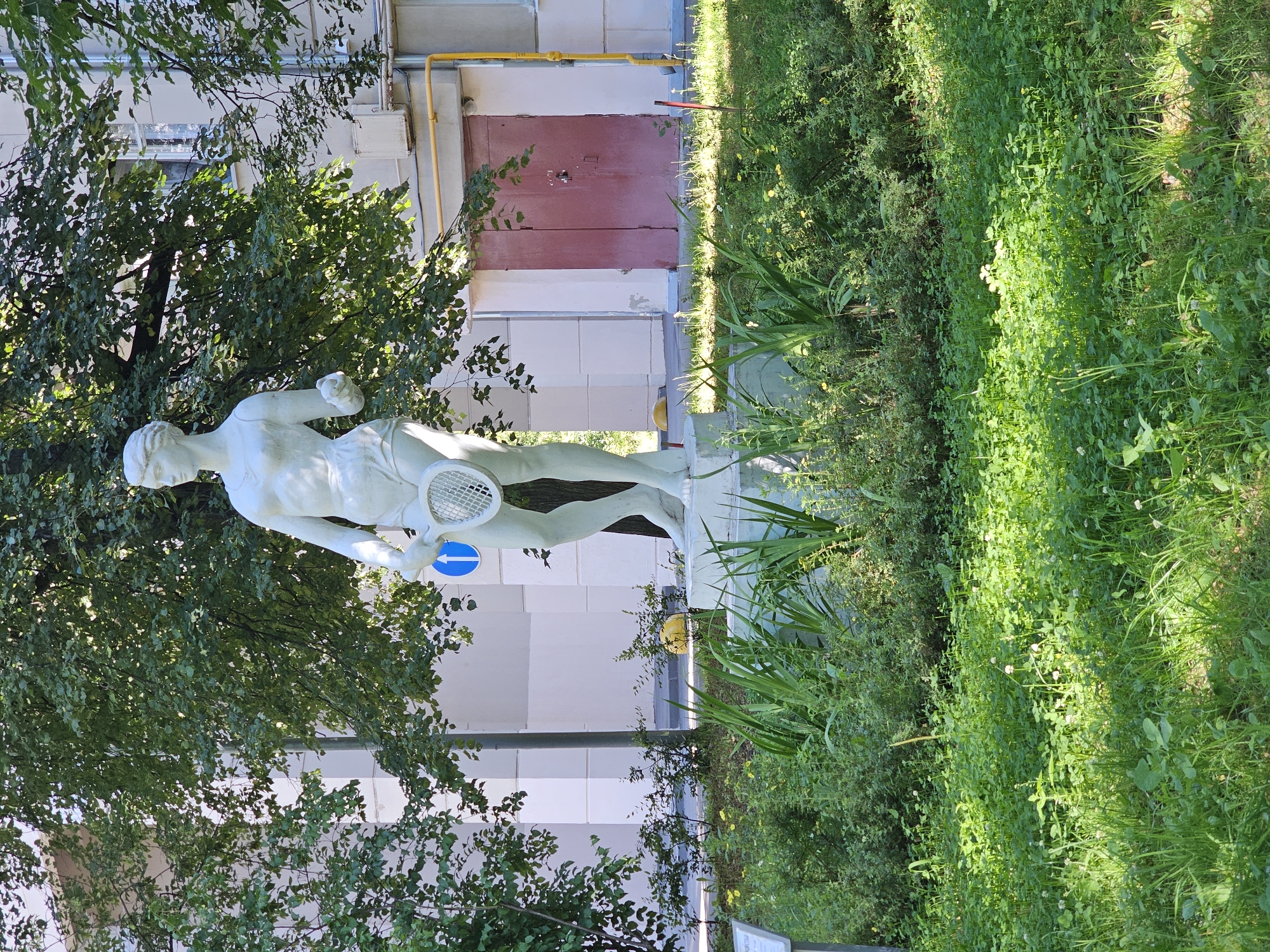 In Khimki, Russia, a white stucco statue of a woman in athletic attire, holding a tennis racket, stands prominently in a grassy area in front of a building. The statue is positioned on a raised concrete pedestal, surrounded by lush green foliage. Behind the statue, the building features a closed, reddish-brown door, windows, and a yellow pipe running vertically along its side. Trees with dense green leaves partially obscure the building and provide shade. The ground in the foreground is covered with short green grass, interspersed with small white and yellow flowers. A blue sign with a white arrow is attached to a pole near the statue. The scene is bathed in sunlight, creating distinct shadows.