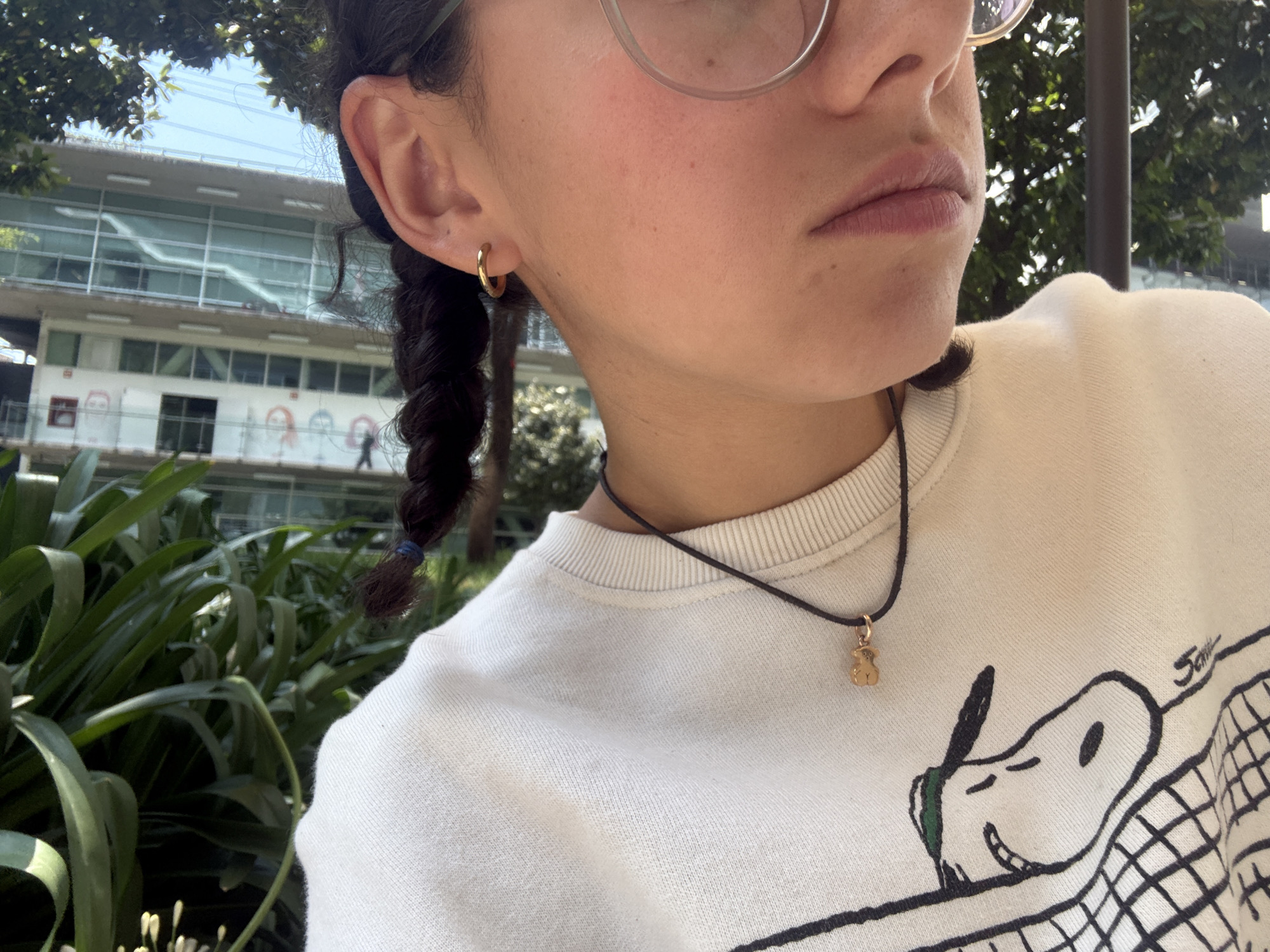 The image captures a close-up, outdoor portrait of a person's right profile and upper body, set against a vibrant green and urban background in what appears to be Mexico City. The subject has dark, braided hair secured with a blue elastic, visible behind their ear. They wear clear-framed glasses and a small, golden hoop earring in their right ear. A black cord necklace with a tiny golden bear pendant rests around their neck.
The person is wearing a light-colored, possibly cream or white, crew-neck sweatshirt. A prominent black line drawing of the character Snoopy from Peanuts is visible on the front of the sweatshirt, smiling behind what appears to be a tennis net. The word "Snoopy" is written above the character. The subject's expression is neutral, with their lips slightly parted.
In the background, lush green foliage dominates the foreground and mid-ground, suggesting a natural or landscaped area. Behind the greenery, a modern, multi-story building with numerous windows is visible under a clear, bright sky. The lower level of the building features colorful, cartoon-like figures or murals, including what looks like a rainbow pattern, adorning its facade. Indistinct figures of people can be seen inside or around the building. The bright light indicates a sunny daytime setting.