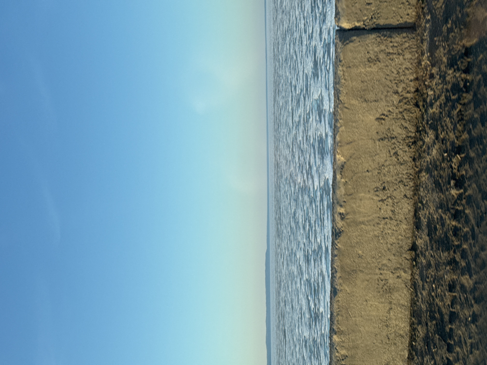 The image shows a body of water, possibly a river or lake, with gentle waves and a light blue hue, bordering a sandy or earthen bank. The sky above is a clear, light blue color. The photograph appears to be taken at the water's edge, with a focus on the boundary between the water and the land. There is no evidence of people, actions, or interactions in the provided image. It is plausible that the location is Chaikovsky, Russia, given the description, but there is no independent confirmation of this from the image itself.
