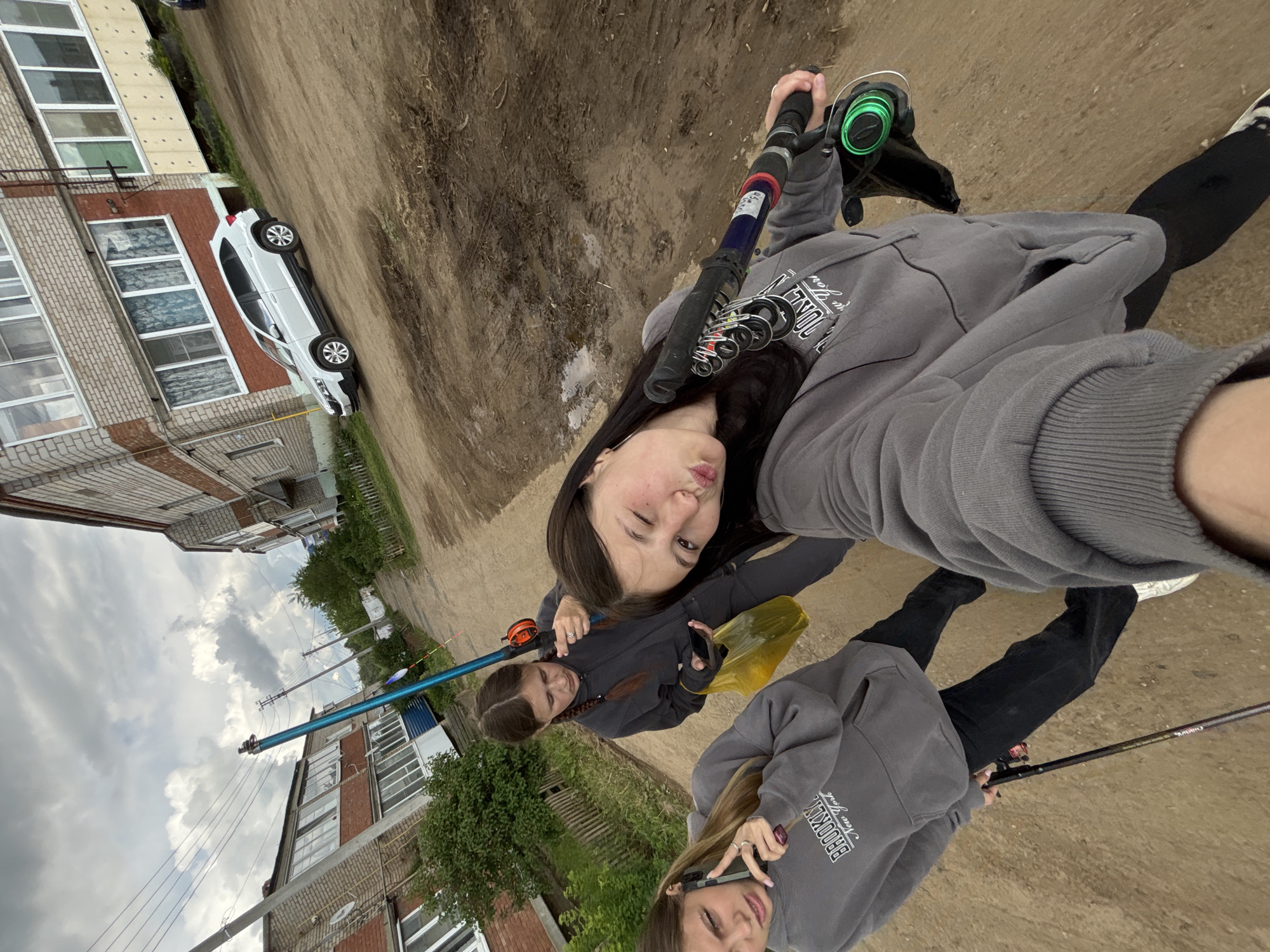 Three young women are taking a selfie on a dirt road in Фоки, Russia. They are holding fishing rods. Buildings are visible in the background. A white car is parked near one of the buildings. The sky is cloudy.