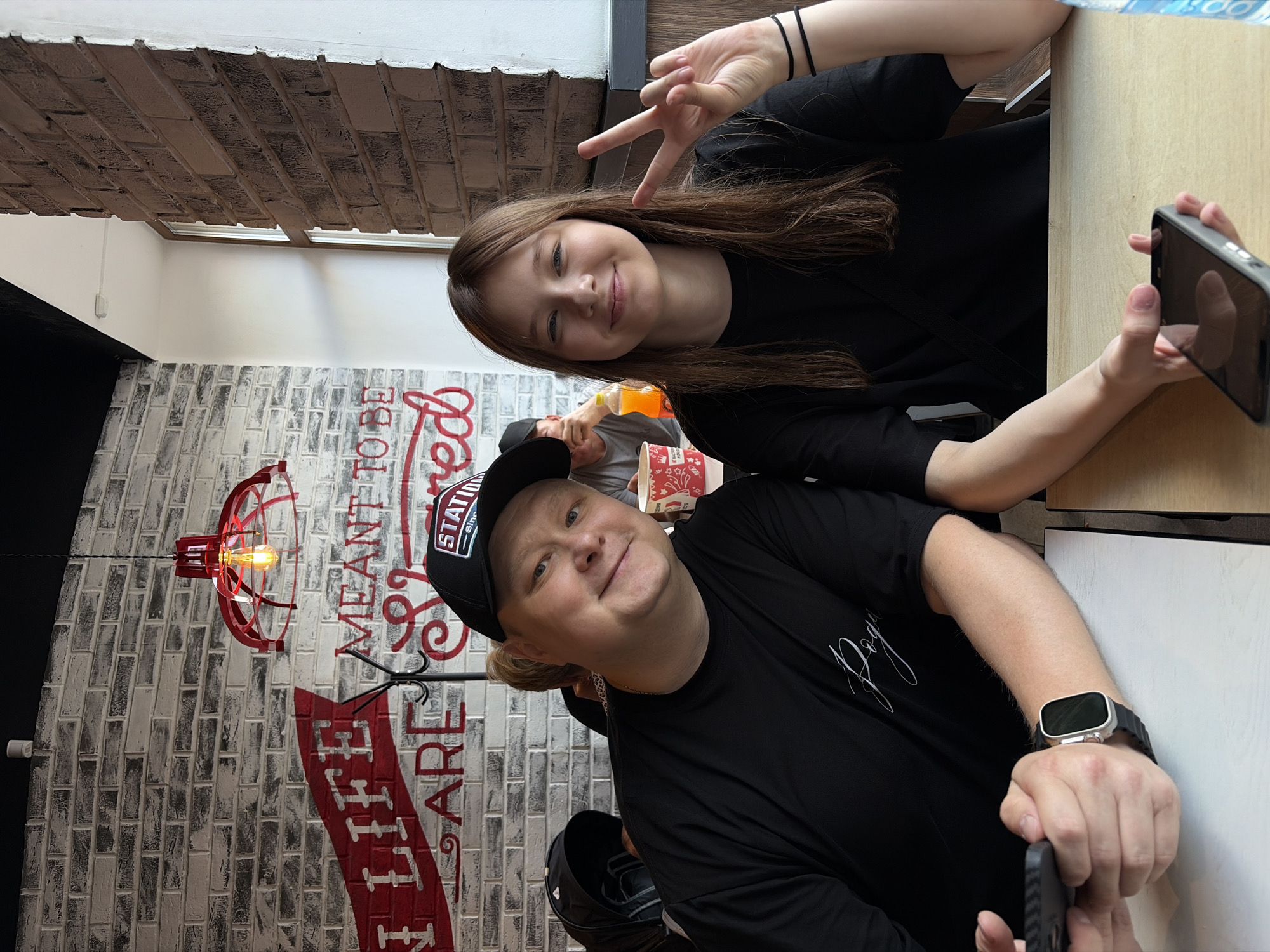 A medium shot shows a man and a young girl seated at a table in a brightly lit restaurant. The man, on the left, is wearing a black t-shirt and a black baseball cap. He is smiling and holding a smartphone in his left hand. A smartwatch is visible on his right wrist. The girl, on the right, is wearing a black t-shirt and has long brown hair. She is smiling and making a peace sign with her right hand. She is holding a smartphone in her left hand. Behind them, a brick wall with red and white lettering is visible. A red light fixture hangs from the ceiling. In the background, other patrons and restaurant decor can be seen. The overall impression is one of a casual outing. The location is a cafe in Chaikovsky, Russia.