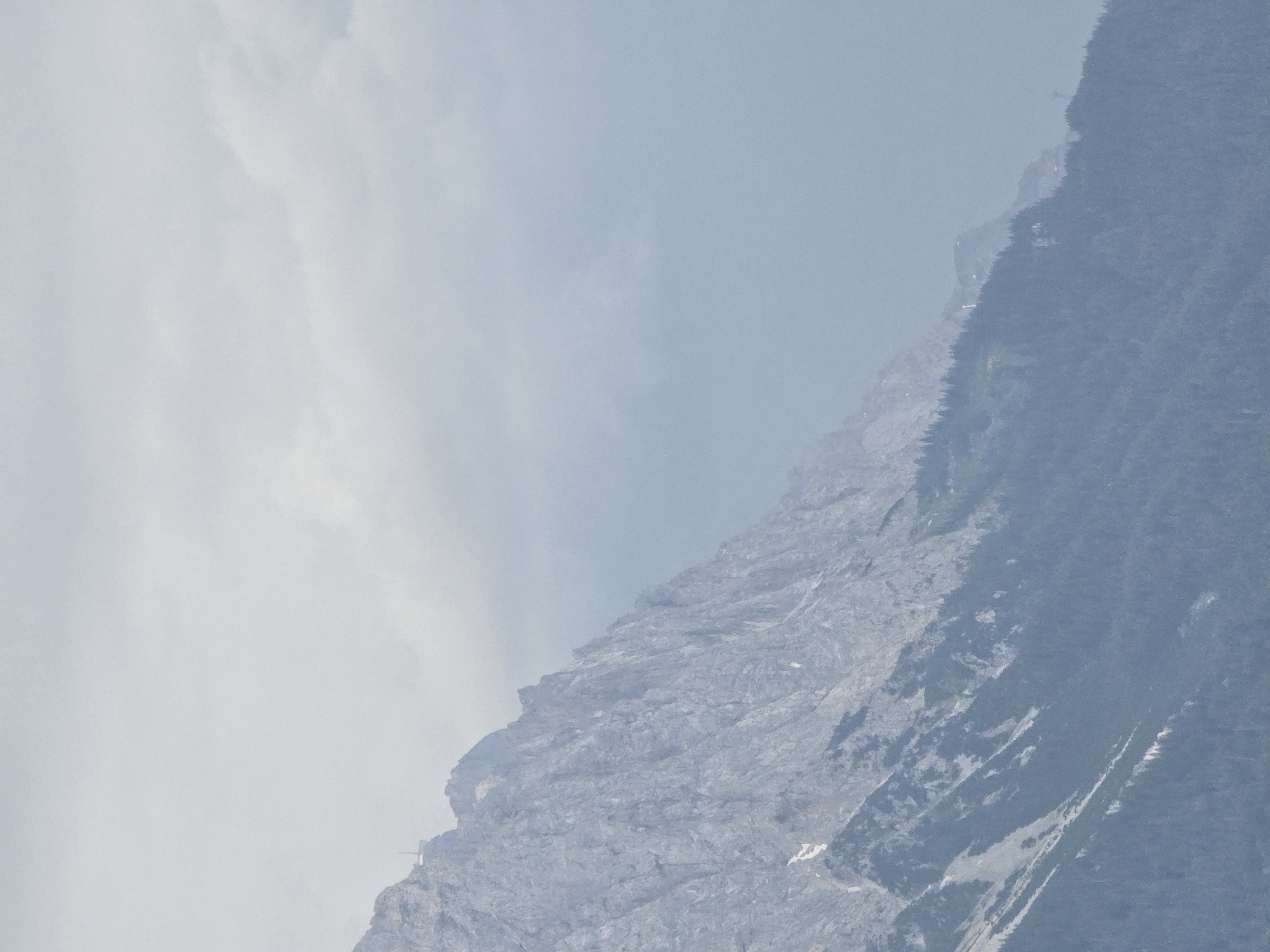 The image depicts a misty, mountainous landscape. The dominant subject is a steep, rocky mountain face that occupies the right and lower portions of the frame. The mountain is partially shrouded in a light haze or fog, which softens its features and creates a sense of depth. The texture of the rock is rough and varied, with visible striations and outcrops.
On the left side of the image, the sky is a pale, hazy blue, suggesting a cloudy or overcast day. Wispy clouds are visible, blending into the atmosphere. The transition between the sky and the mountain is gradual due to the mist, making it difficult to discern a sharp horizon line.
In the upper right corner of the image, a small, bright red object, possibly a distant aircraft or a structure on a mountain peak, is barely visible against the muted background. Towards the middle of the mountain slope, a few small white patches might indicate snow or patches of sunlight breaking through the mist. A thin line of dark green, likely trees or vegetation, is visible along a lower ridge of the mountain, adding a touch of color to the otherwise muted palette.
The scene evokes a feeling of natural grandeur and perhaps isolation. The lighting is diffuse, indicating it is likely daytime, though the specific time is not clearly discernible. The location is identified as Garmisch-Partenkirchen, Germany, a region known for its alpine scenery, which aligns with the visual elements of the image. There is no visible text in the image.