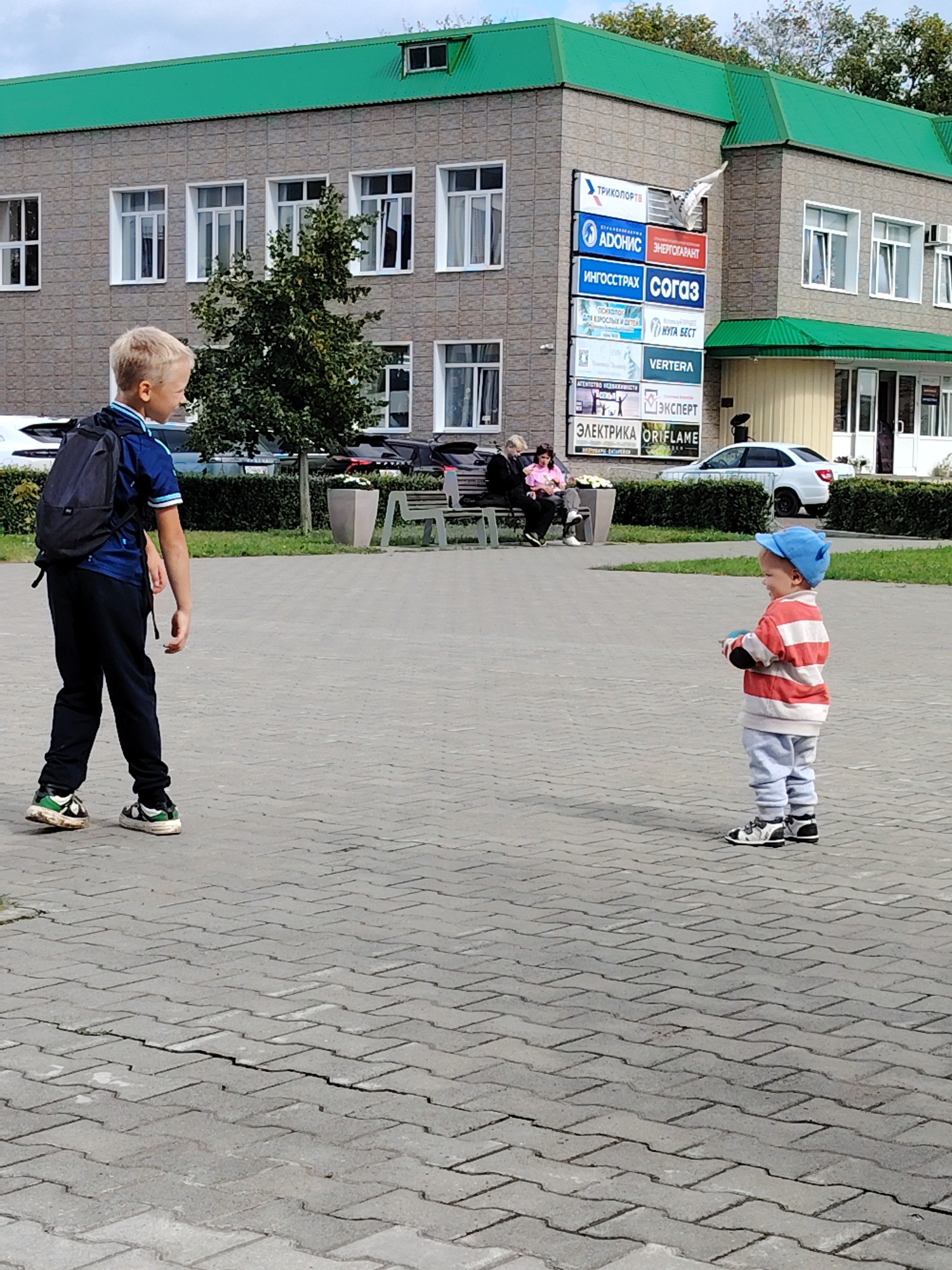 This is an outdoor, eye-level, full-shot photograph of two boys in a paved public area in front of a two-story building with a green roof in Chaikovsky, Russia. The boy on the left is older, wearing a blue polo shirt with white trim, black pants, and a black backpack. He is facing right, looking away from the camera. The younger boy on the right is wearing a blue baseball cap, a striped red and white long-sleeved shirt, grey pants, and black and white sneakers. He is facing left, looking towards the older boy, and holding a blue object. In the background, there are several parked cars and a building with multiple windows. A sign with various logos and text in Russian is visible on the side of the building. The ground is paved with grey interlocking bricks, some of which are cracked. A bench and planters are also visible in the background. The sky is overcast.