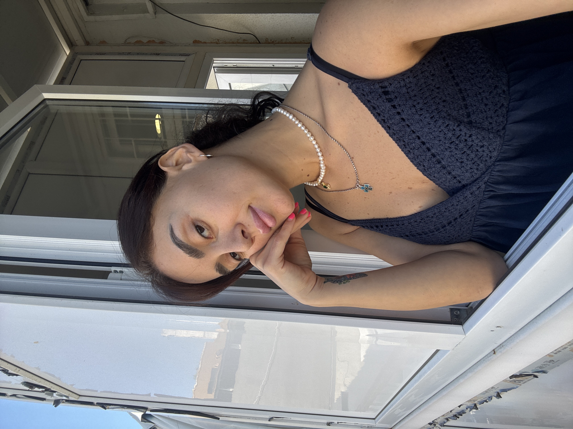 A young woman with dark hair and light skin leans her head on her hand, looking towards the camera. She wears a dark blue dress with a crochet-like texture on the bodice and a pearl necklace with a cross pendant. Her left arm is visible, showing a tattoo on her forearm. She is positioned near an open window with white frames. The background is indistinct, with reflections visible in the window. The lighting suggests it is daytime. The location is identified as Tbilisi, Georgia.