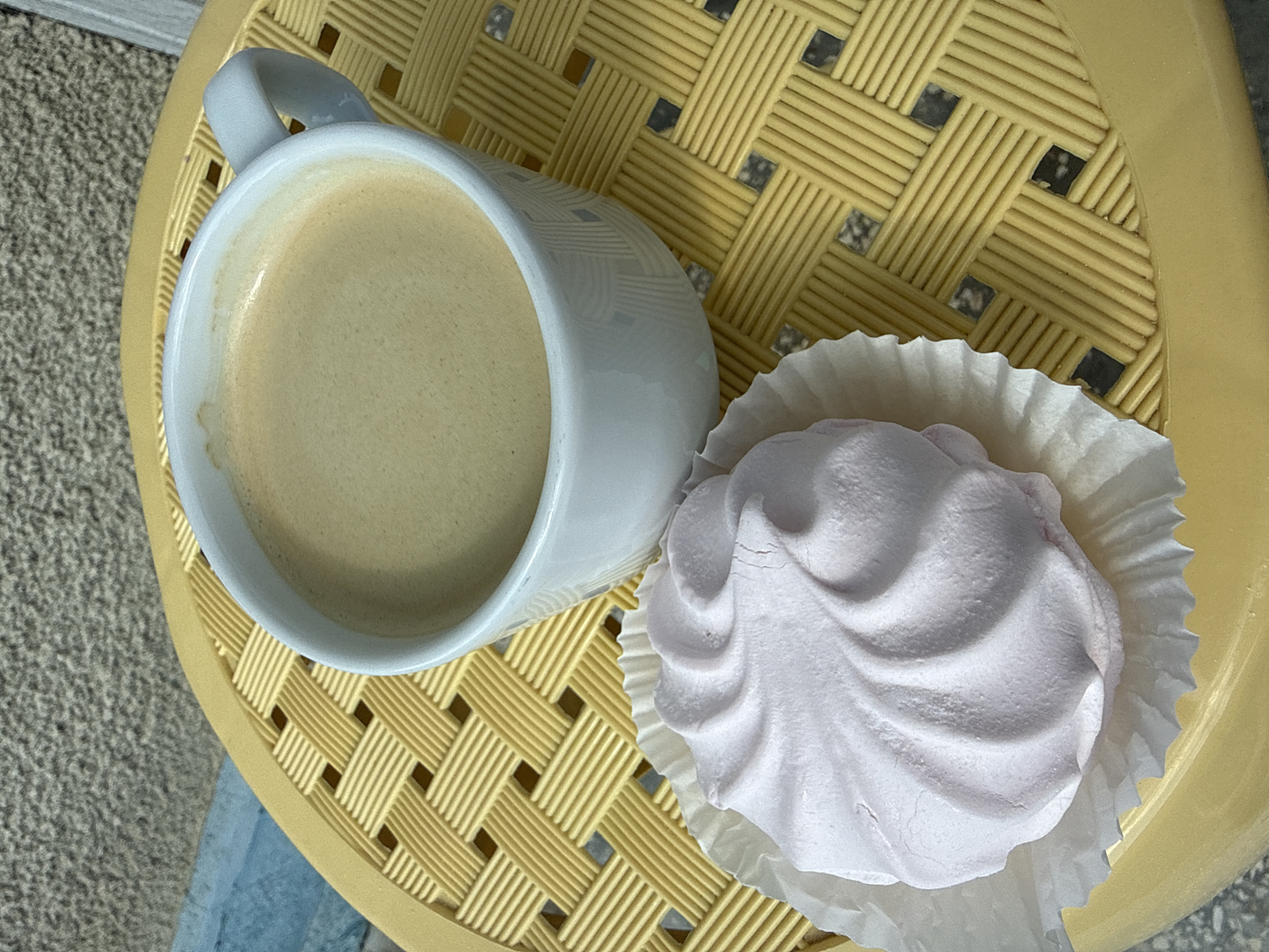 An overhead, high-angle medium shot shows a white ceramic mug filled with frothy, light brown coffee sitting on a yellow woven plastic tabletop. To the left of the mug is a pink, swirled marshmallow in a white paper liner. The background consists of a textured gray wall and a blue surface. The scene appears to be outdoors.