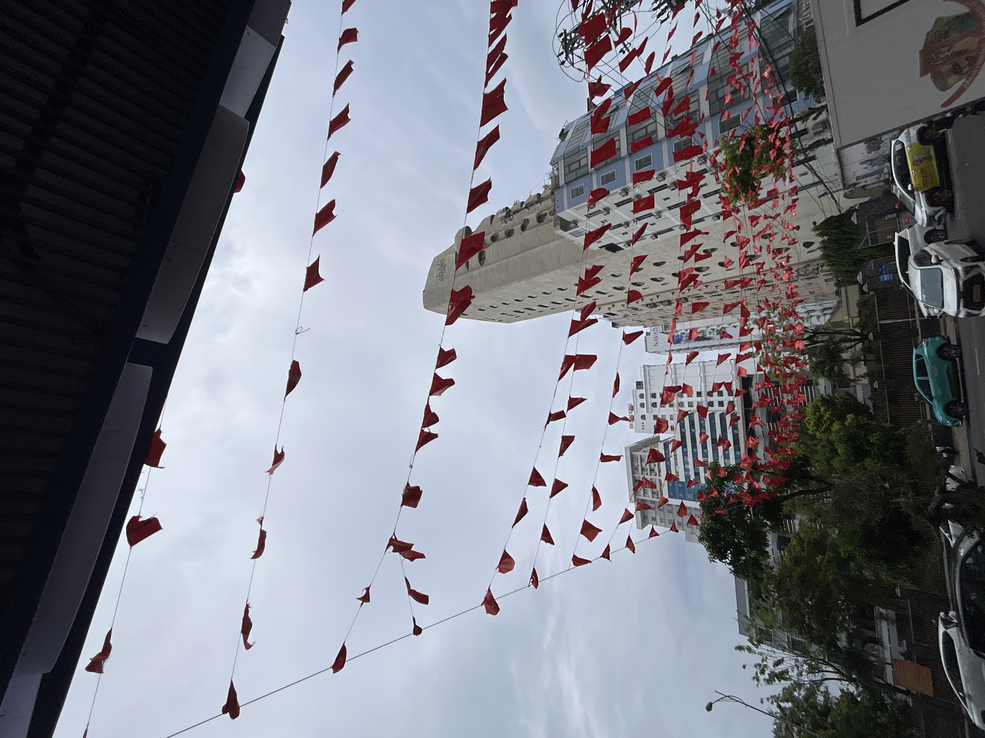 The photograph, taken in Bac Nha Trang Ward, Vietnam, is shot from a low angle, looking upwards. Red flags are strung across the street, partially obscuring the view of the buildings and the sky. The sky is overcast. On the left side of the frame, a dark, overhanging structure suggests the edge of a building. In the background, various buildings are visible, one being notably taller and white with multiple windows. On the right side, parked cars are visible, including a yellow taxi. Trees with dense foliage are present amidst the buildings. The overall scene depicts a street decorated with red flags, likely for a local celebration or event.