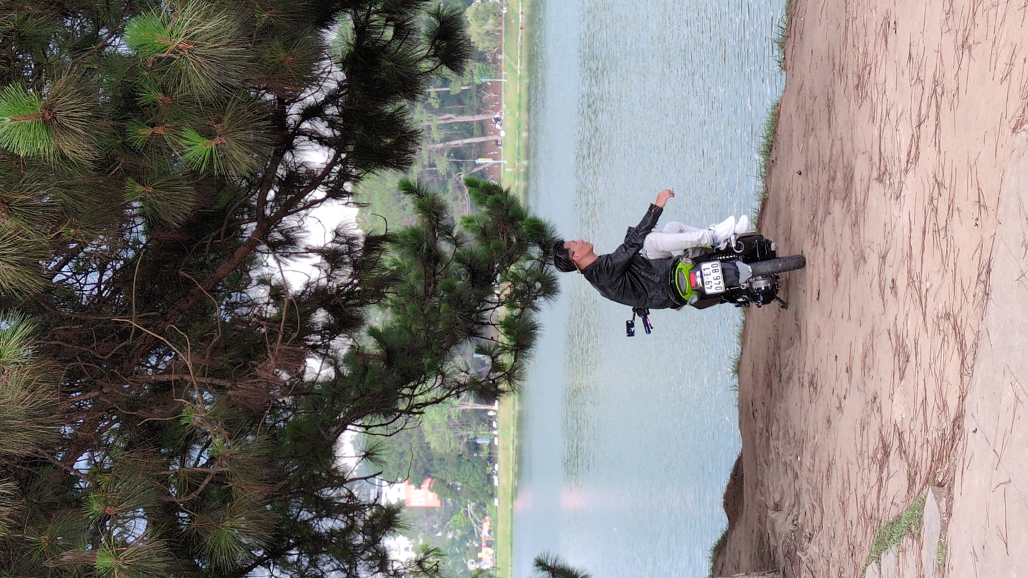 A scenic photo reveals a figure in a black jacket and white pants, reclined on a motorcycle positioned on a sandy verge near a large body of water; the vehicle's license plate is legible. Overhanging foliage, primarily pine, frames the image's left side, while across the water, hints of urban development can be discerned, indicating the photo was taken at Da Lat Lake, within the city of Da Lat, Vietnam.