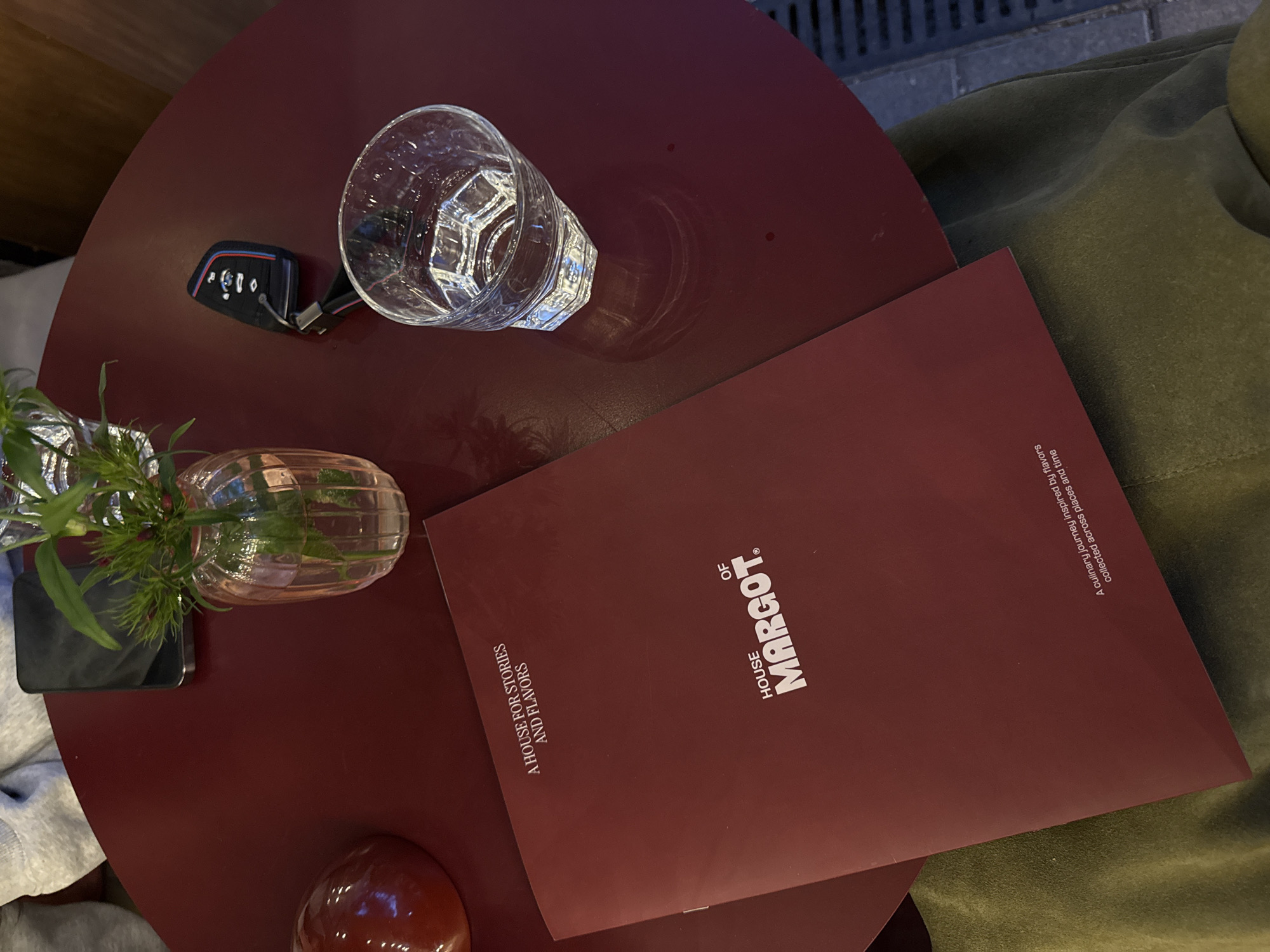 A high-angle shot captures a round, reddish-brown table setting, potentially within a restaurant or cafe in Yerevan, Armenia. The table's surface accommodates several items: a partially filled glass of water with light refracting through it, a BMW car key, a small vase containing water and fresh sprigs of greenery, a smartphone, and a menu from "House of Margot." The menu, in a matching reddish-brown hue, displays white typography. Additionally, the periphery of the frame shows part of an olive green jacket and some stone flooring near a building.