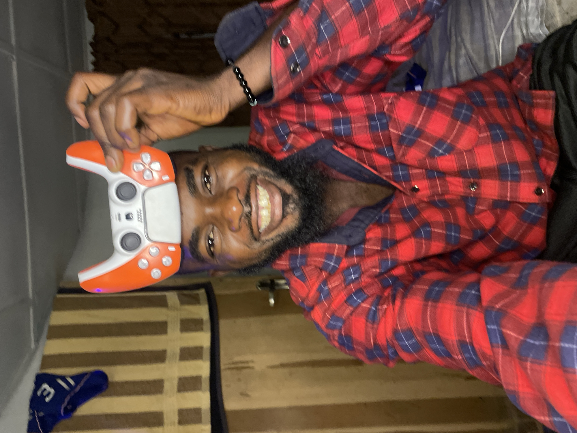 A smiling Black man with a beard, wearing a red and blue plaid shirt, is posing for a selfie. He is holding a white and orange game controller on top of his head. In the background, there is a striped curtain and a door handle. There is also a blue sock with a white number "3" on it hanging on the left side of the image. The environment appears to be a domestic setting, possibly a bedroom, within the city of Jos, Nigeria.