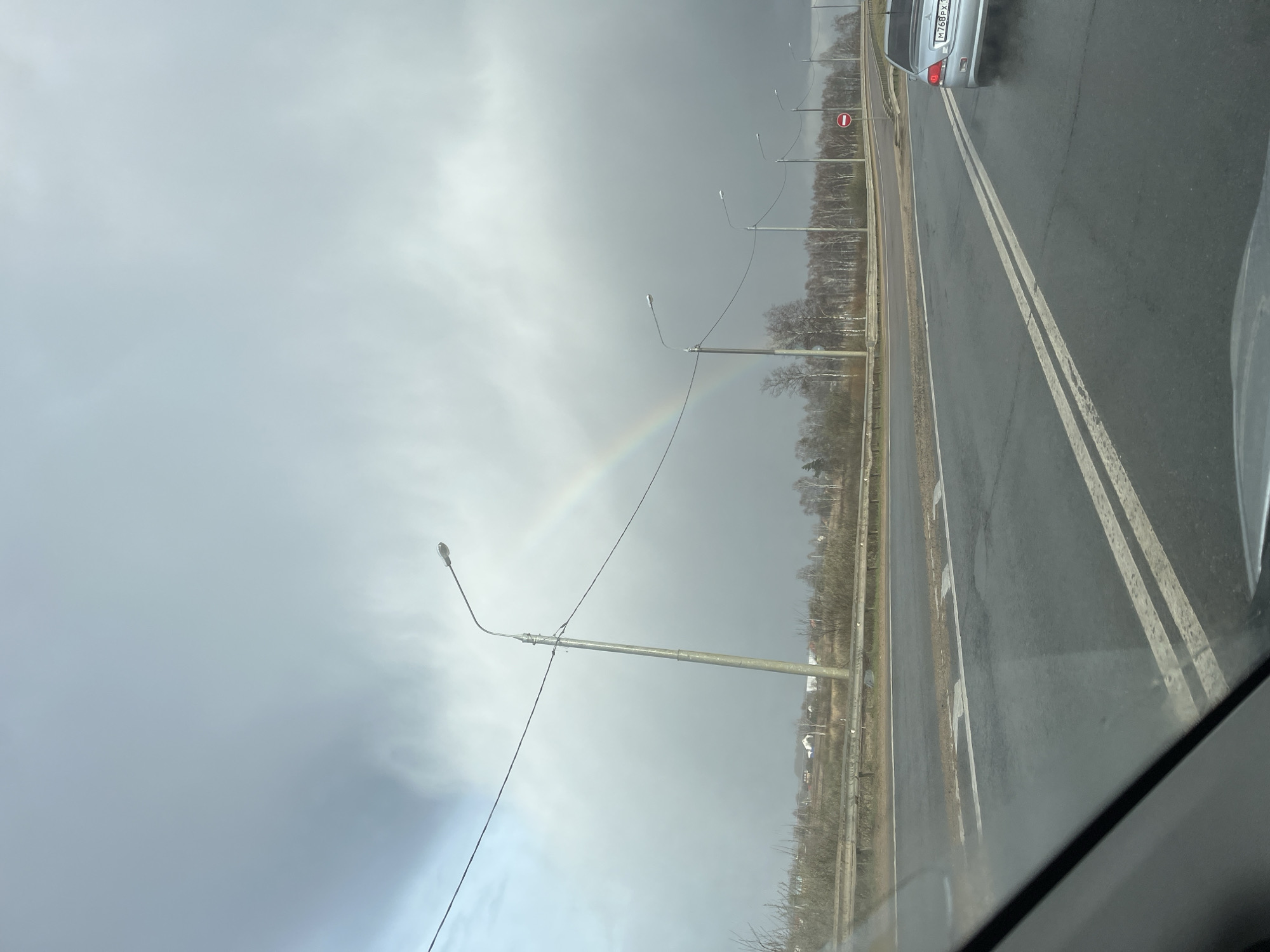 The image shows a roadside view from within a vehicle, possibly in Ivashevo, Russia. A faint rainbow arches across a gray sky. The road is wet with double white lines marking lanes. Streetlights line the road's edge with bare trees in the background. A silver car is visible in the adjacent lane. The scene is overcast.