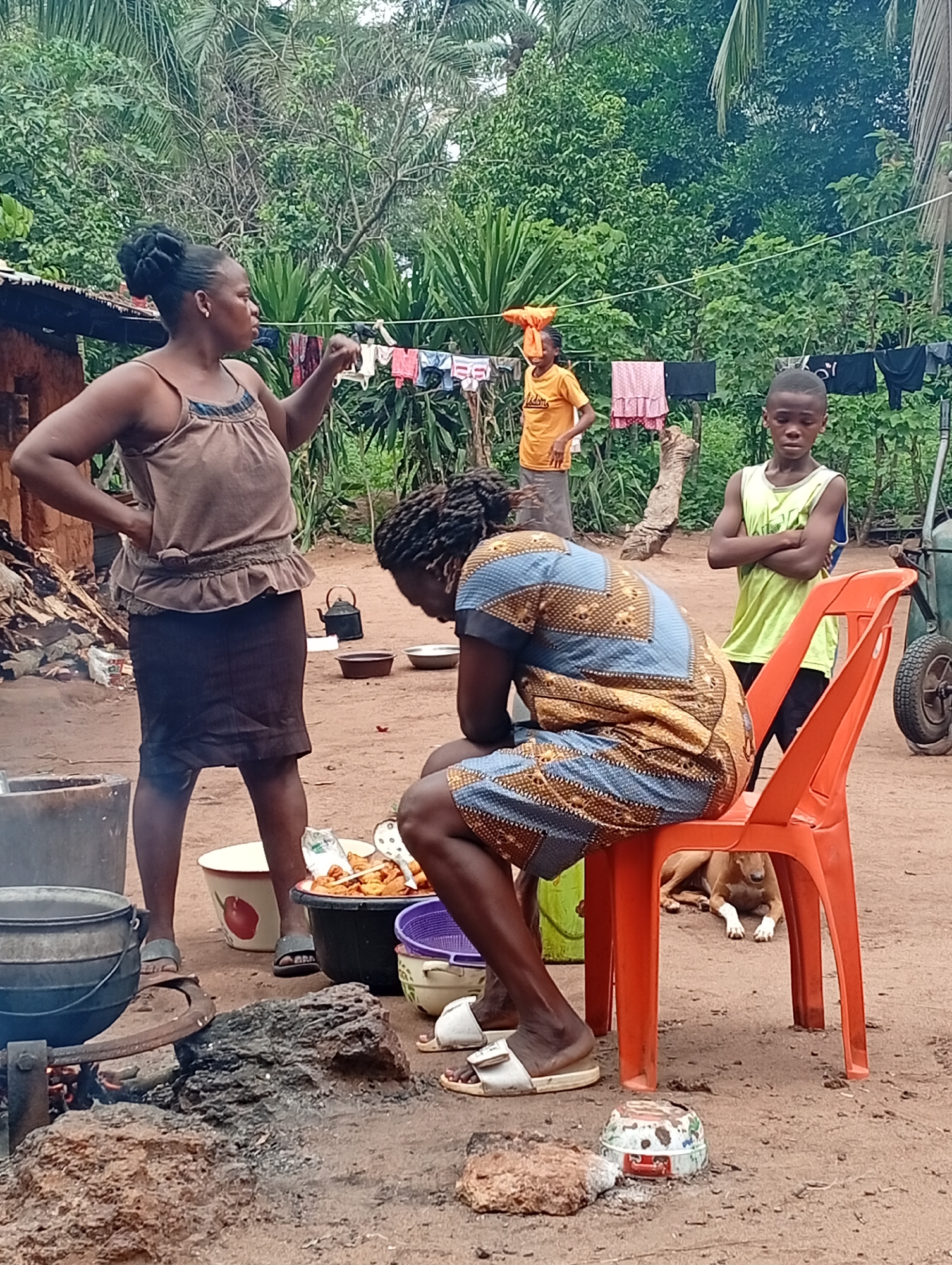 In Adda, Nigeria, a woman in a brown sleeveless top and a dark skirt gestures with her right hand while speaking to another person. She is standing in a dusty yard surrounded by lush green foliage. In the foreground, a woman in a blue and gold patterned dress sits on an orange plastic chair. Her head is bowed, and she appears to be resting. A young boy in a light green sleeveless shirt stands with his arms crossed, looking towards the camera. In the background, a younger child in an orange t-shirt stands near a clothesline with various garments hanging. Several pots and bowls are visible around the yard, suggesting cooking activities. A brown dog lies on the ground beneath the orange chair. The overall scene depicts a domestic outdoor setting with elements of daily life and possibly food preparation.