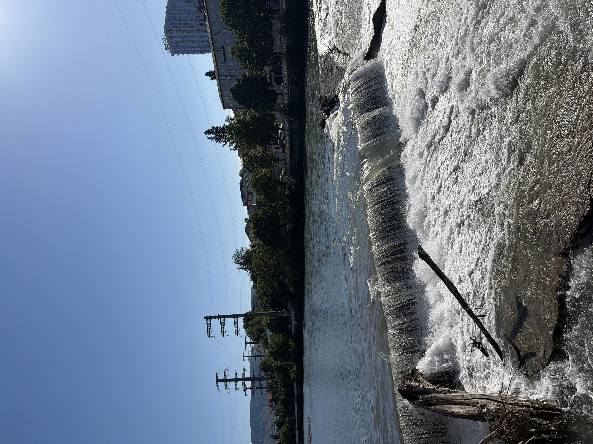 This media file presents a sunlit landscape view of a wide river flowing through an urban and natural setting in Sochi, Russia.
The river dominates the foreground and midground, featuring a significant weir or waterfall on the right side where water vigorously cascades, creating abundant white foam and turbulent flow. Several large pieces of driftwood, including a prominent log, are caught in the rushing water near the waterfall.
The left bank of the river showcases a blend of human development and lush greenery. Numerous green trees line the bank, interspersed with multi-story buildings. One tall, light-colored building is visible with many windows and unreadable text, likely Cyrillic, on its facade (appears to be "БИЗНЕС ЦЕНТР"). Further along this bank, several large metal power line towers with multiple overhead wires stand against the sky, indicating electrical infrastructure. Distant hills or mountains, partially obscured by trees, form the backdrop.
The sky is a clear, bright blue, suggesting a sunny day. The sun casts strong reflections on the water, illuminating the foamy cascades. No people are visible, and no specific activity is taking place beyond the natural flow of the river. The overall scene depicts a dynamic natural waterway integrated into an urban landscape under clear weather conditions.