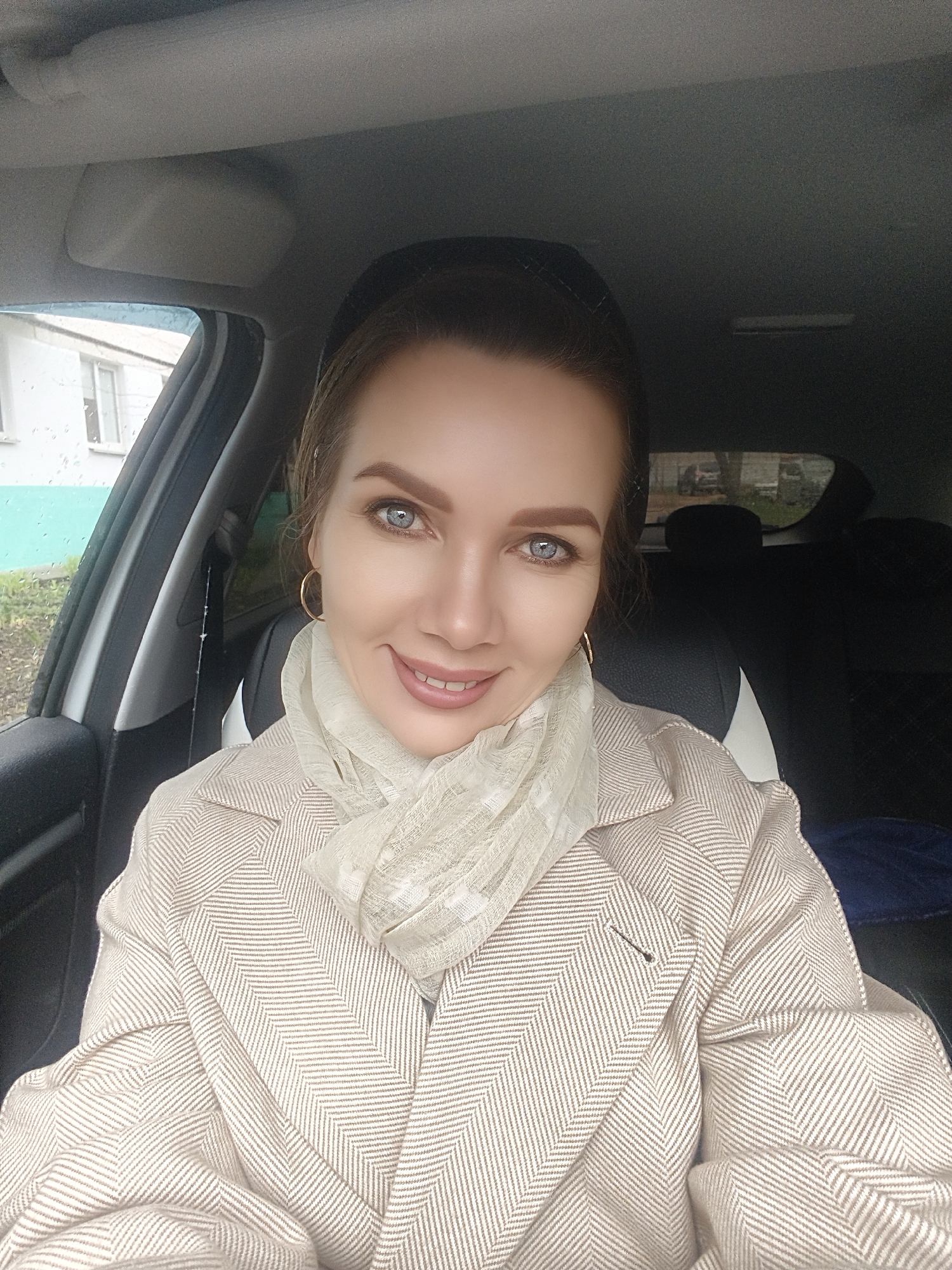 A selfie captures a woman from the chest up, seated inside a car. The woman is the primary subject, featuring light skin, prominent blue eyes, and light brown hair styled back. She exhibits a gentle, pleasant smile, looking directly into the camera. She is dressed in a light-colored, possibly beige or off-white, coat with a distinct herringbone pattern, and a matching or complementary sheer scarf wrapped around her neck. Gold hoop earrings are visible on both ears.
The setting is inside a car, likely the driver's seat, with parts of the vehicle's interior such as the headliner, dashboard, and a car seat visible. Outside the car's side window, numerous raindrops indicate recent or ongoing rain and an overcast day. Through the wet window, a light-colored building with dark windows and some outdoor foliage can be seen, suggesting a residential or suburban backdrop. The overall lighting is soft, highlighting her face against the slightly darker car interior. No text is visible in the image.