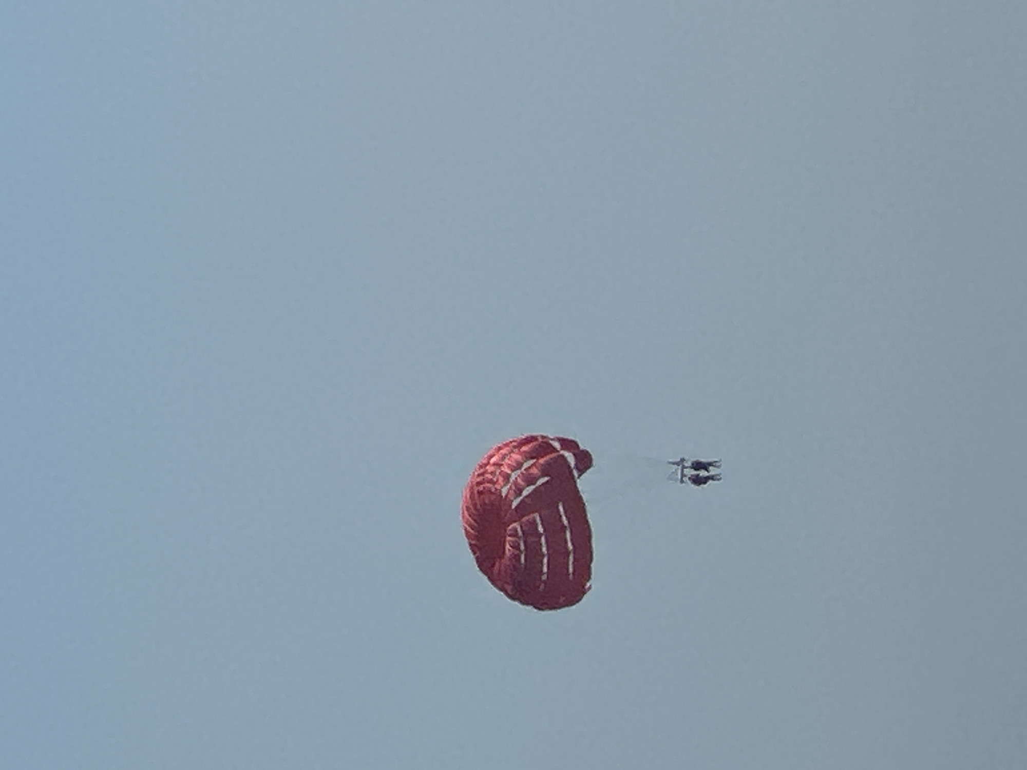 A red and white parasail floats in a clear blue sky over Boreti, Montenegro. Two people are visible below the parasail, suspended by ropes. The parasail is inflated and appears to be in good condition. The sky is clear and the weather is calm. The scene depicts a leisure activity taking place outdoors.