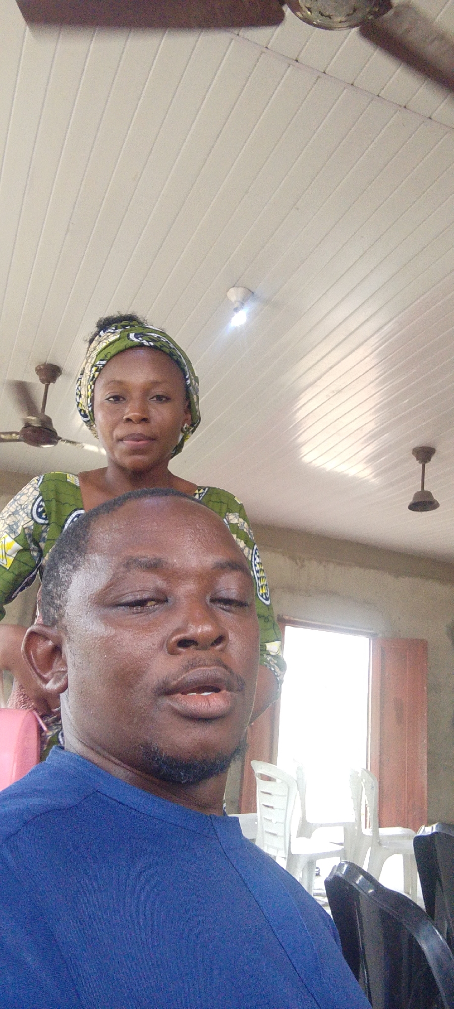 A selfie of a man in a blue shirt is captured from a low angle. Behind him stands a woman in a green and white patterned top and headwrap. The location appears to be indoors, evidenced by the ceiling with multiple ceiling fans and a fluorescent light fixture. There are white plastic chairs in the background, and a window is visible. The location is Lokoja, Nigeria.