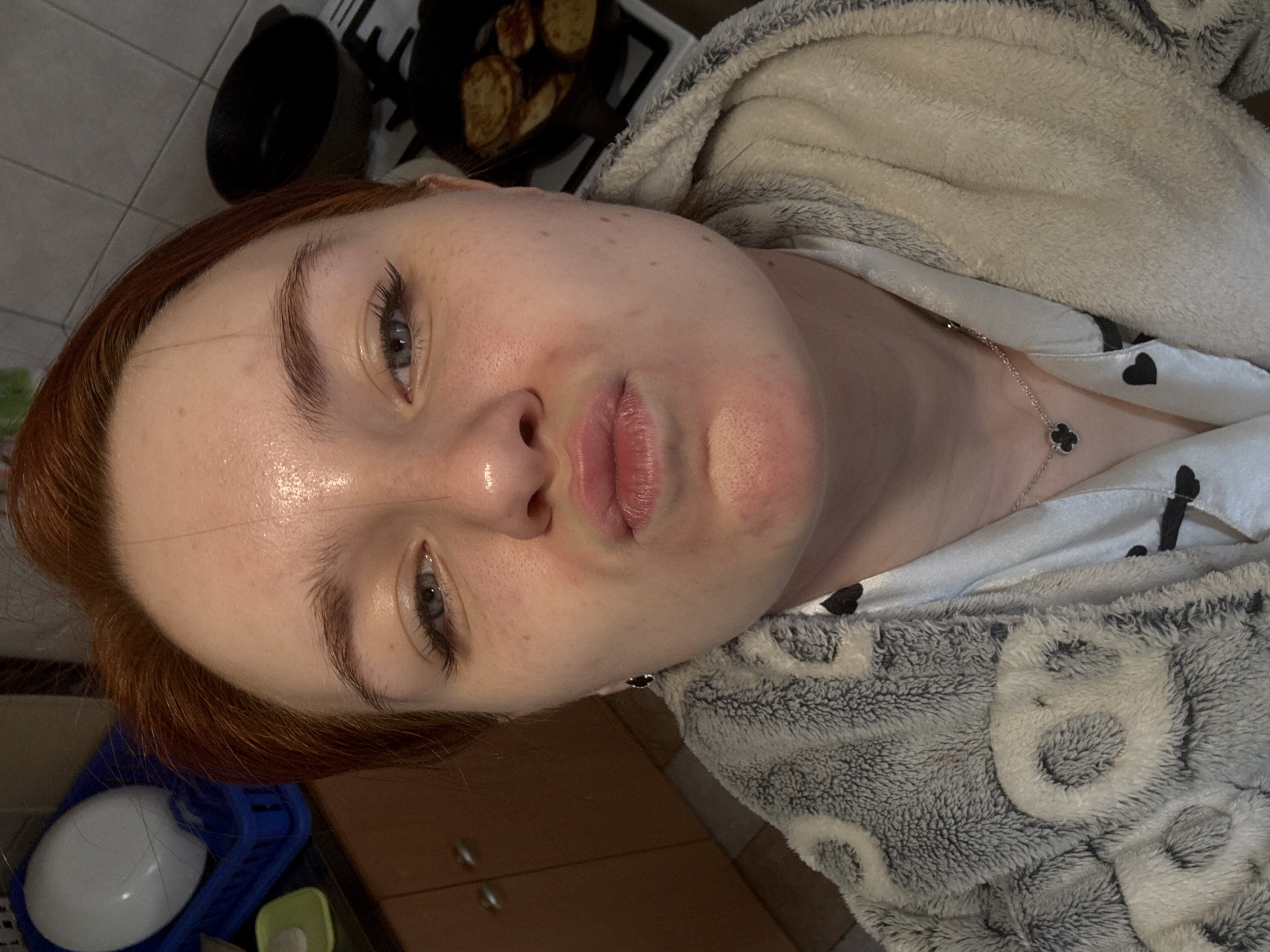 A close-up, tilted shot of a woman's face, neck, and partially visible upper body. The woman has fair skin, reddish-brown hair, light-colored eyes, and visible freckles. Her lips are slightly pursed. She is wearing a light-colored pajama top with black heart patterns and a silver necklace with a black clover pendant. A grey and white fuzzy hooded robe is partially visible over her pajamas.
In the background, there is a tiled floor, a wooden cabinet, and a blue dish rack containing a white bowl. A black pan with food items inside sits on a white and black patterned surface. The setting appears to be indoors, possibly a kitchen. There is no information indicating the city of Sinyavino, Russia in the image.