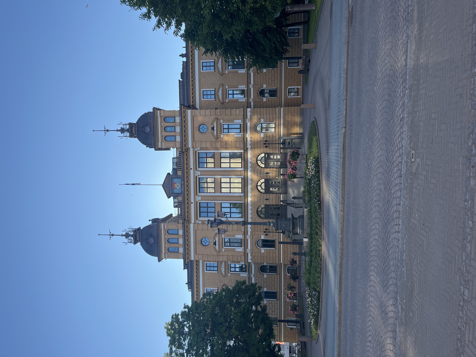 The image shows a large, ornate, tan-colored building with two distinct towers topped with dark domes and intricate spires, against a clear blue sky. The building features numerous windows, some arched and some rectangular, and decorative architectural elements. In front of the building is a circular garden area with flowering plants and a lamppost. A paved walkway and road lead up to the entrance of the building, which has a set of stairs. Two large trees flank the building. The scene appears to be a public square or plaza in the city of Oulu, Finland. The building is likely the Oulu City Hall. There are no people visible in the image. The lighting suggests it is daytime, likely morning or late afternoon given the long shadows cast.