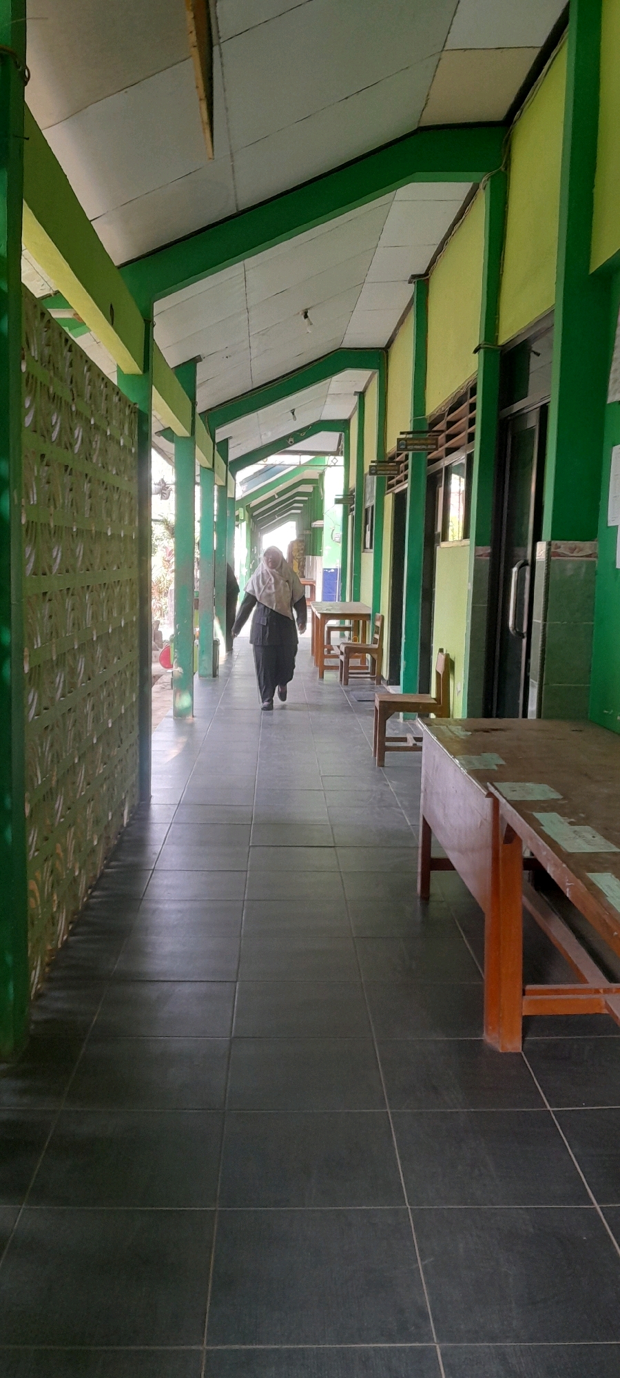 The image shows a woman in a hijab and dark clothing walking down a tiled hallway in what appears to be a school or institutional building. The hallway has a green and yellow exterior with a white tiled ceiling supported by green pillars. There are several wooden tables and chairs lining the right side of the hallway. A decorative green screen is visible on the left side of the hallway. The scene is likely outdoors or semi-outdoors, given the natural light and the presence of plants visible through an opening on the left. The location is specified as Tahunan, Indonesia.