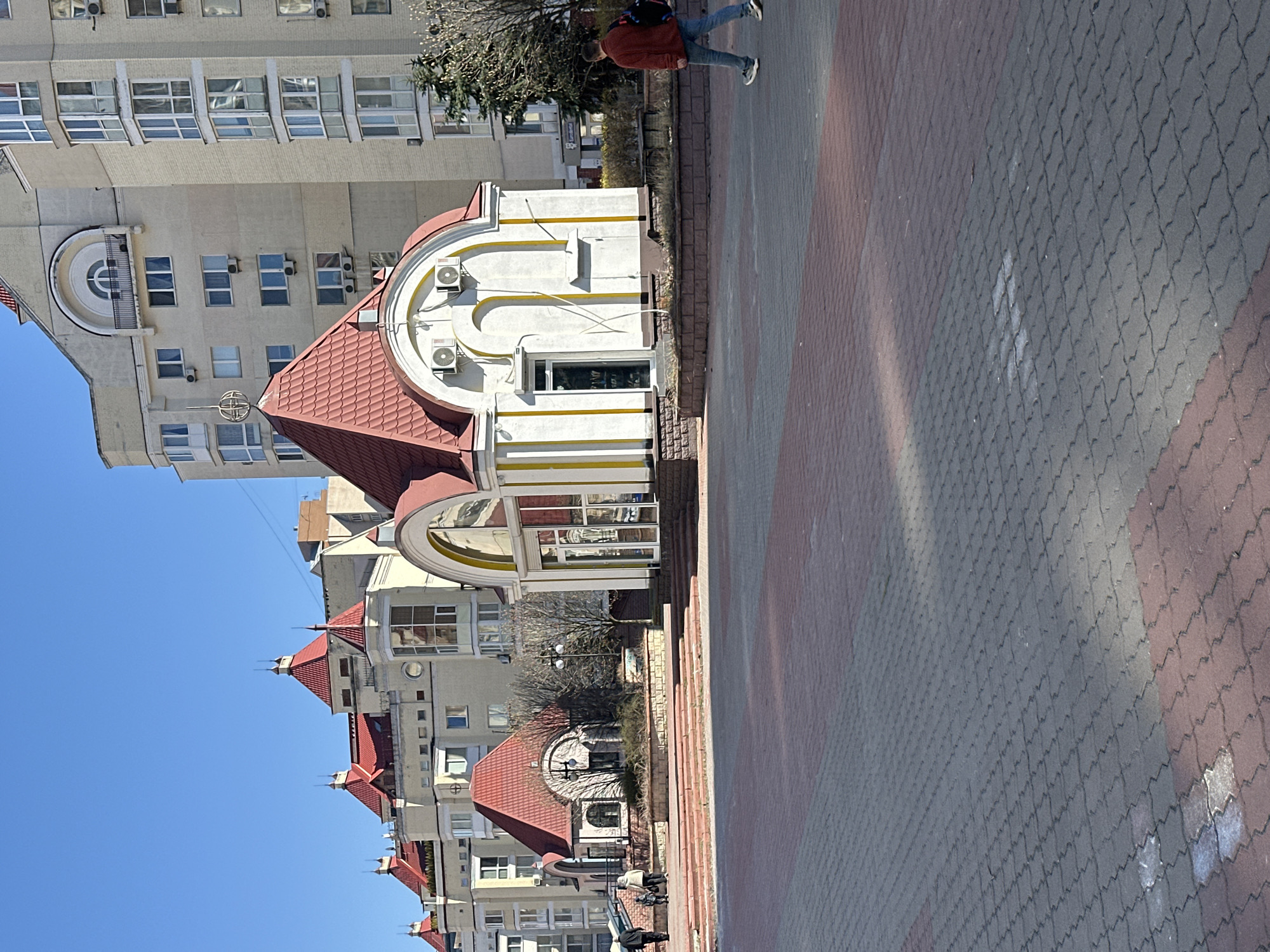 The image depicts a cityscape in Kyiv, Ukraine, showcasing a blend of architectural styles. In the foreground is a paved walkway with a distinctive brick pattern in hues of gray and reddish-brown. A person wearing a red jacket and jeans can be seen walking along the sidewalk.
Adjacent to the walkway is a small, ornate white building with a red-tiled roof. The building is adorned with yellow accents and appears to be a kiosk or small shop. Behind this structure, there is a row of residential buildings with similar red-tiled roofs. These buildings are multistoried, featuring numerous windows and architectural details like arched balconies.
In the backdrop, a taller building is visible, characterized by its beige facade and repetitive pattern of windows. The sky is clear and blue, indicating a sunny day. The environment is urban, with a mix of commercial and residential structures. The presence of air conditioning units on the buildings suggests a modern adaptation to the climate.
