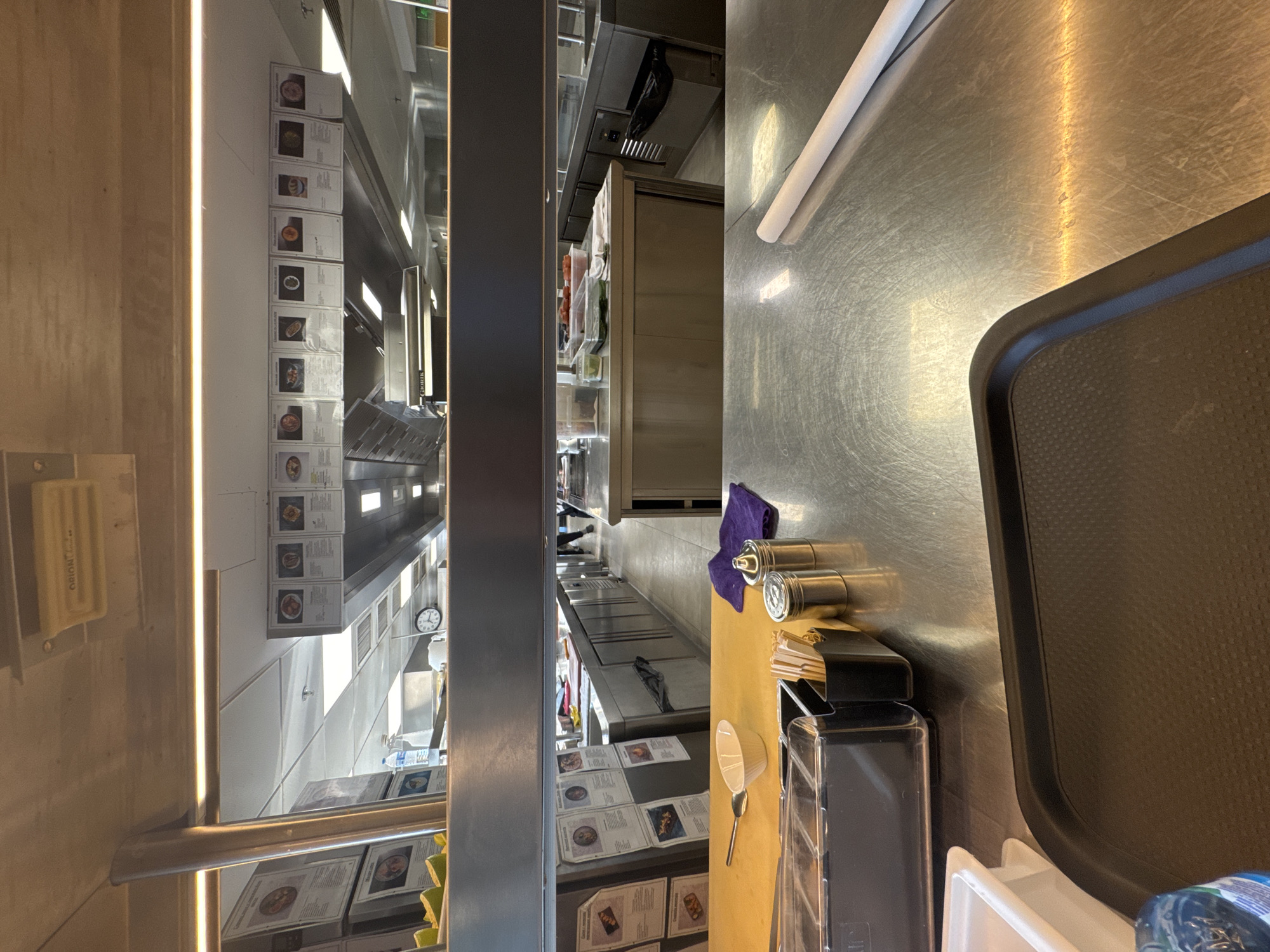 The image depicts a commercial kitchen environment, likely a cafeteria or restaurant serving line, observed from a serving area. The foreground is dominated by a stainless-steel countertop with a black serving tray. To the left, a white plastic container is visible. To the right of the tray is a section of a white paper roll. In the middle ground, there's a yellow-ish serving surface with a small white bowl, a spoon, and a purple napkin. Adjacent to these are two small metal containers, one appearing to be a salt or pepper shaker, and a black organizer filled with cutlery or skewers.
In the background, through a horizontal opening, a wider view of the kitchen is presented. This area features stainless-steel counters, cabinets, and equipment. Several framed menus or informational sheets displaying images of food items are mounted on the wall. A clock with a white face and black hands is visible on the ceiling. Lighting fixtures are integrated into the ceiling. There are some people in the distance, appearing to be staff members, wearing dark clothing. One individual is walking away from the camera. The overall impression is one of a functional food service establishment.
The image does not contain enough information to determine its specific location within Sofia, Bulgaria.