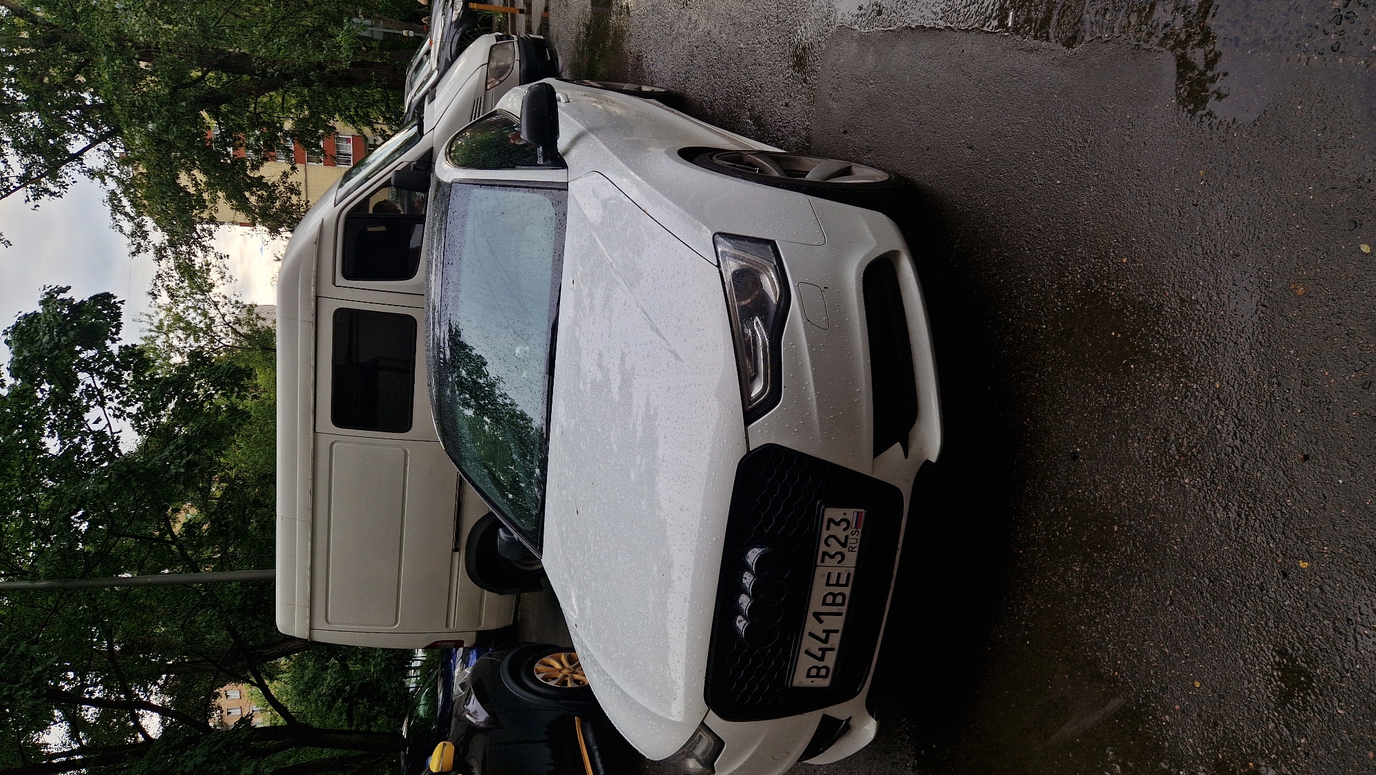 The image shows a white car with a black grille and a Russian license plate that reads "B441BE 323 RUS". The car is parked on a wet asphalt surface, suggesting it might be raining or has recently rained. In the background, there's a white van and trees. A building with windows is visible behind the trees. The scene appears to be an outdoor urban setting, possibly a parking lot or street in Moscow, Russia. The lighting suggests it's daytime, though the sky is overcast. There are no people visible in the image. The focus is on the parked cars and the environment.