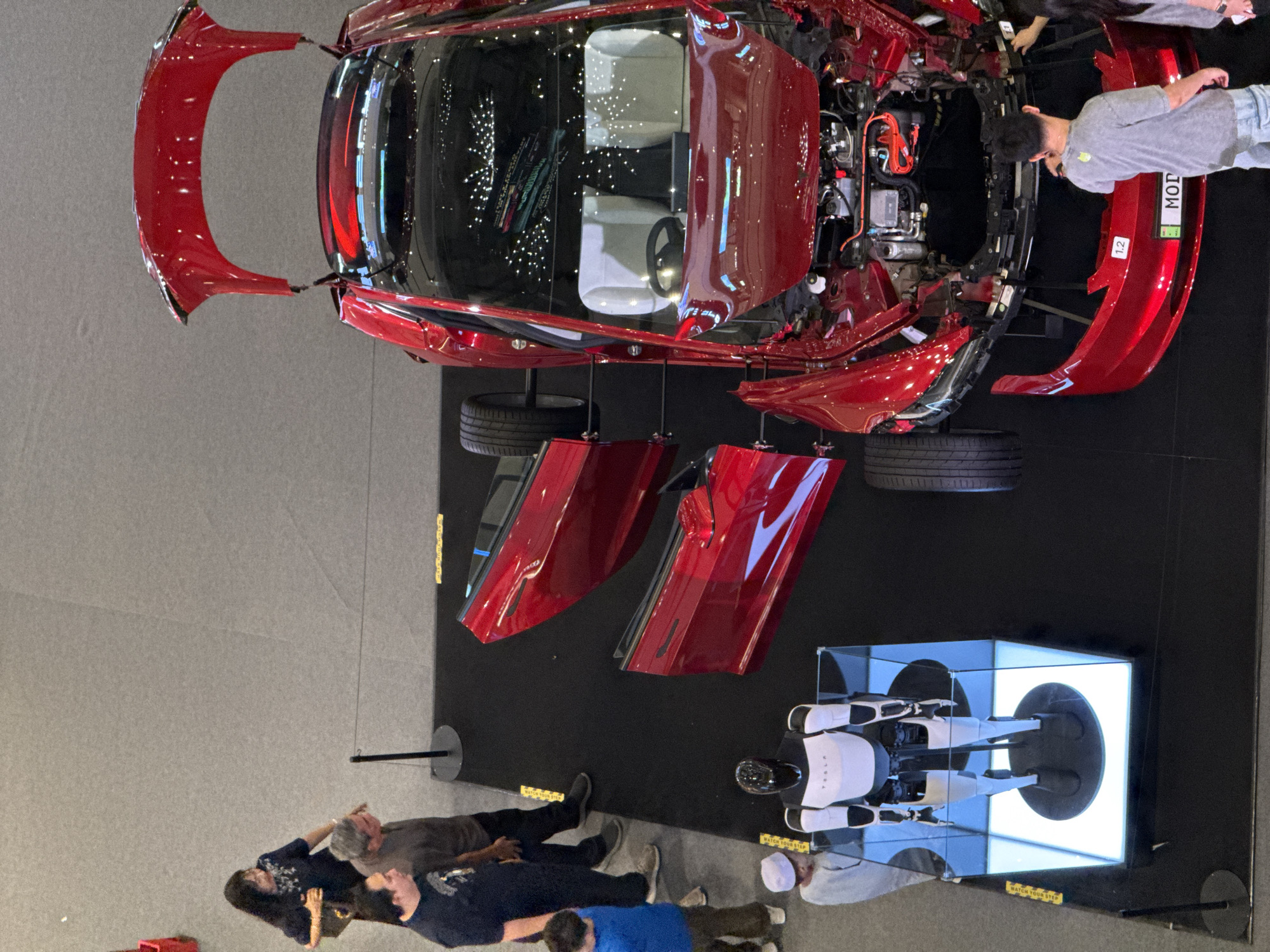 The image shows a disassembled red Tesla car on display in Kuala Lumpur, Malaysia. The car's body parts, including the hood, doors, and bumpers, are separated and suspended above the chassis. People are gathered around the display, observing the car's components. A Tesla Bot model is also part of the exhibit. The environment appears to be an exhibition hall with a dark floor.