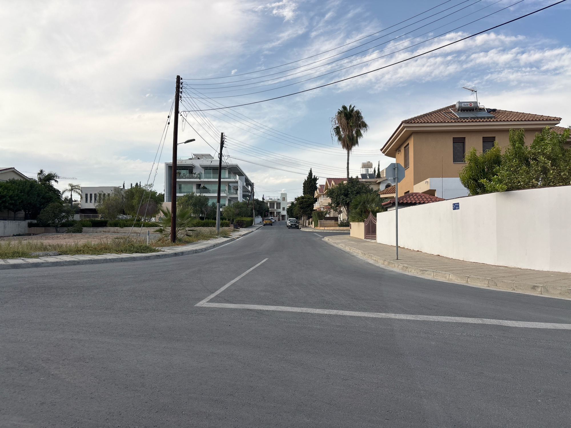The image shows a street in Mesa Geitonia, Cyprus, on a cloudy day. The road is paved and has white lane markings. On the right side of the road, there is a white wall and a house with a tiled roof. There are also trees and power lines visible. On the left side of the road, there are more houses and trees, as well as a tall pole with wires. Two cars are parked on the street in the distance.