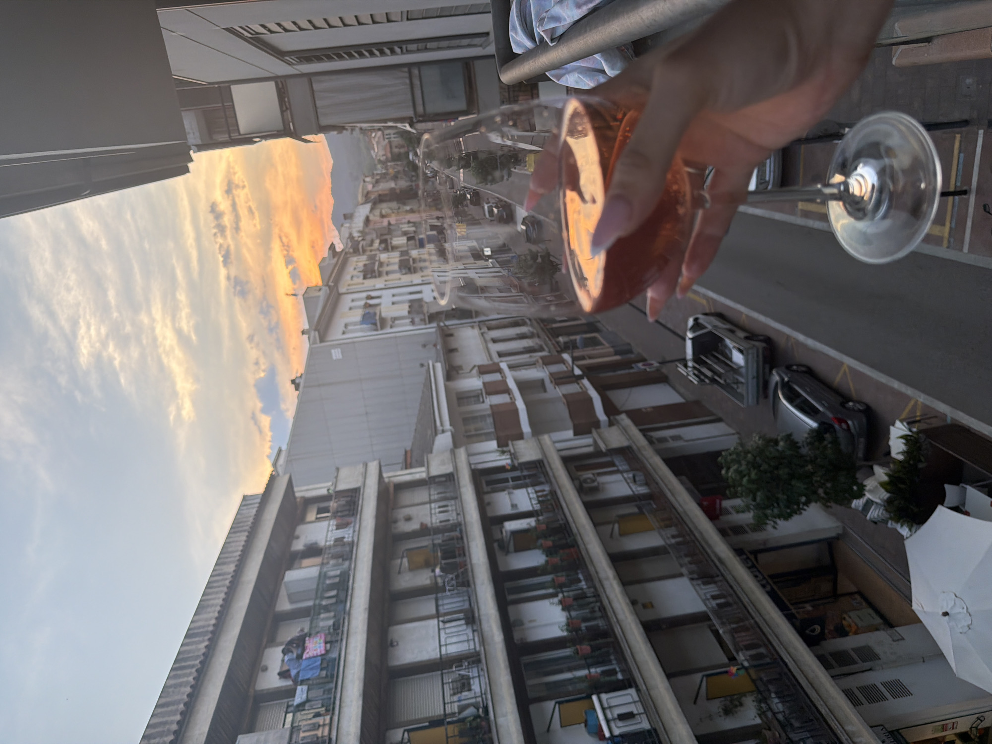 A hand with manicured pink nails holding a wine glass filled with a pink beverage is in the foreground of the image. The glass is reflecting the warm hues of a sunset sky, which is visible over a European-style street scene. The street is lined with multi-story buildings, many with balconies. Laundry is seen hanging on some of the balconies. Cars are parked and driving on the street below. The overall impression is a relaxed evening view from a balcony, likely in a Mediterranean town such as Arbúcies, Spain.